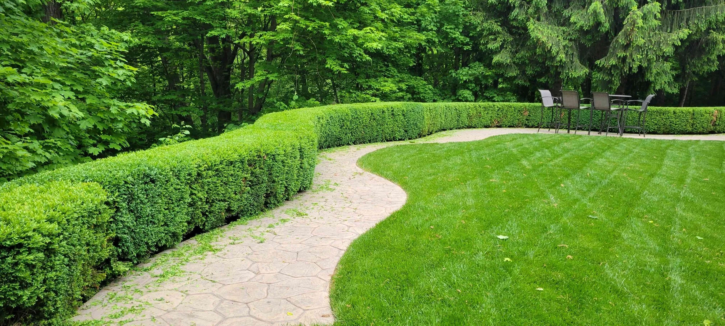 A well-manicured garden with green grass, a curved stone pathway, trimmed hedges, and a set of chairs and a table in the background surrounded by lush trees.