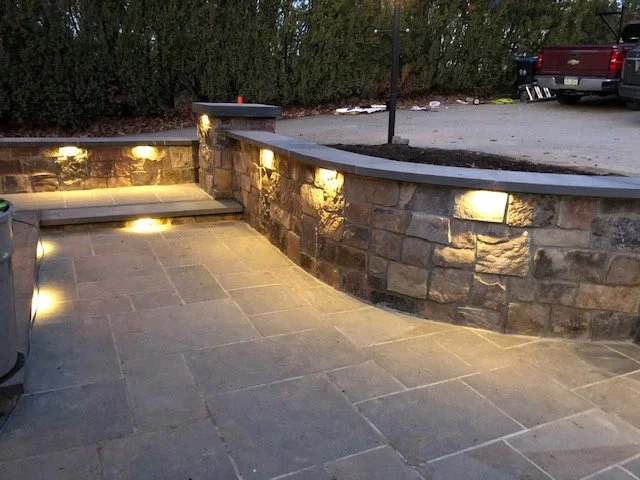 Stone patio with wall lights, surrounded by hedges and a parking area with a red pickup truck.