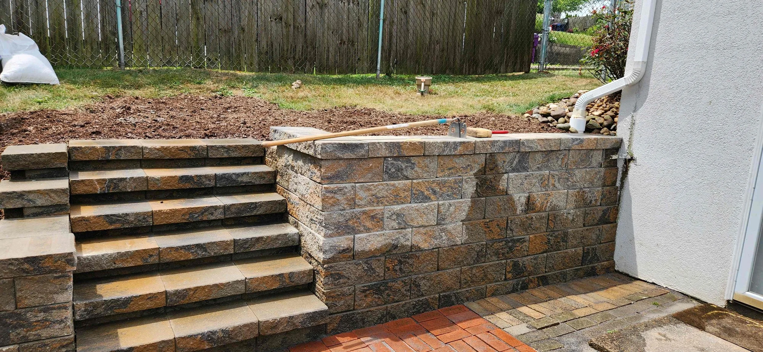Stone steps and retaining wall in a backyard with garden tools on top, surrounded by a grassy area and chain-link fence.
