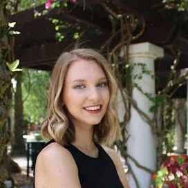 A young woman with wavy blonde hair smiling outdoors near greenery and trees.