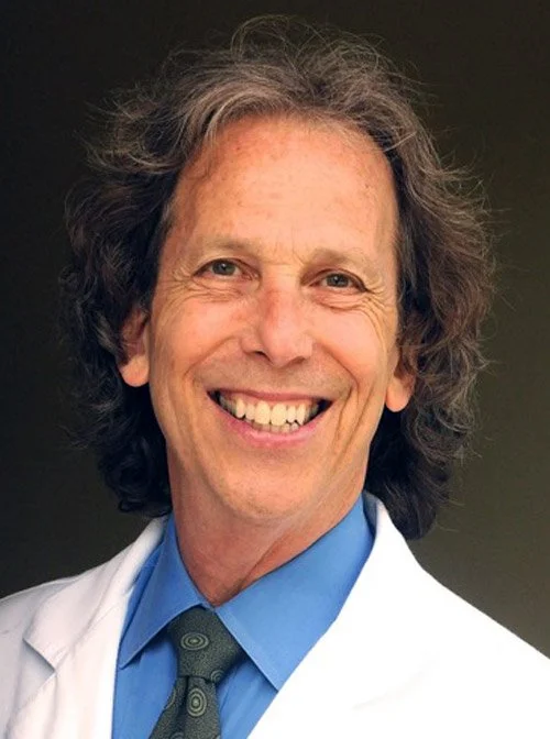A smiling man with curly gray hair, wearing a white lab coat, blue shirt, and patterned tie against a dark background.