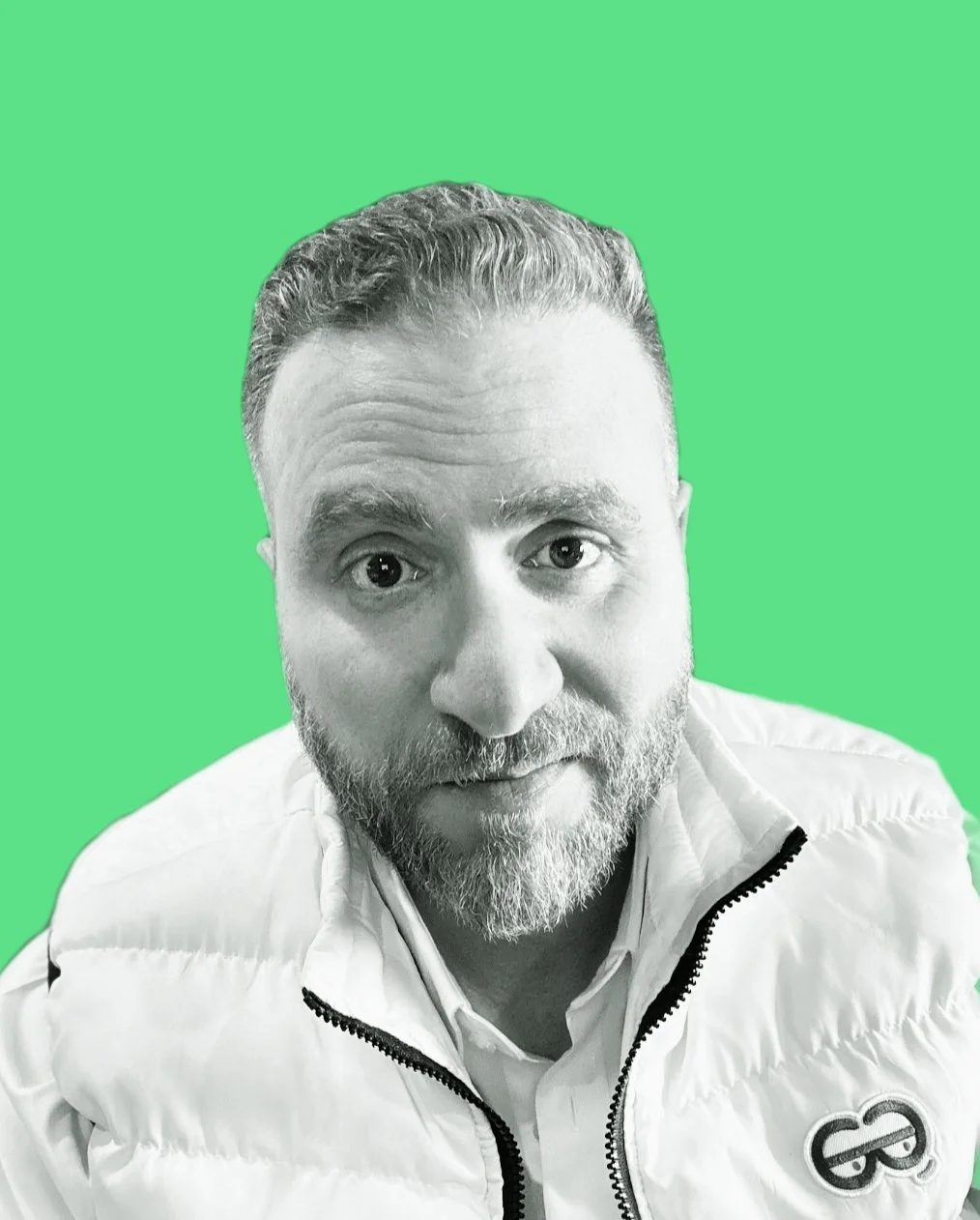 Black and white photo of a man with a beard and mustache, looking directly at the camera, wearing a white jacket with a logo, against a green background.