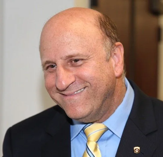 A smiling middle-aged man with a bald head and light skin in a suit, light blue shirt, and yellow striped tie, indoors.