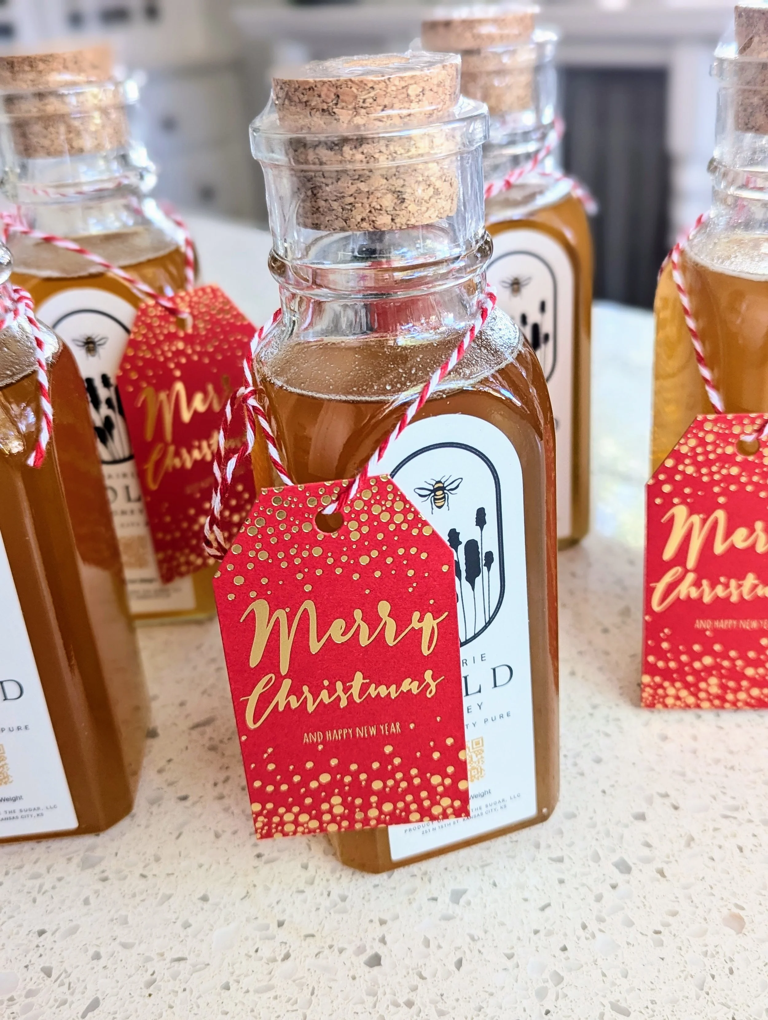 Bottles of honey with cork stoppers, each with a red and white string and a red label that says 'Merry Christmas' and 'And Happy New Year' in gold text, decorated with gold dots.