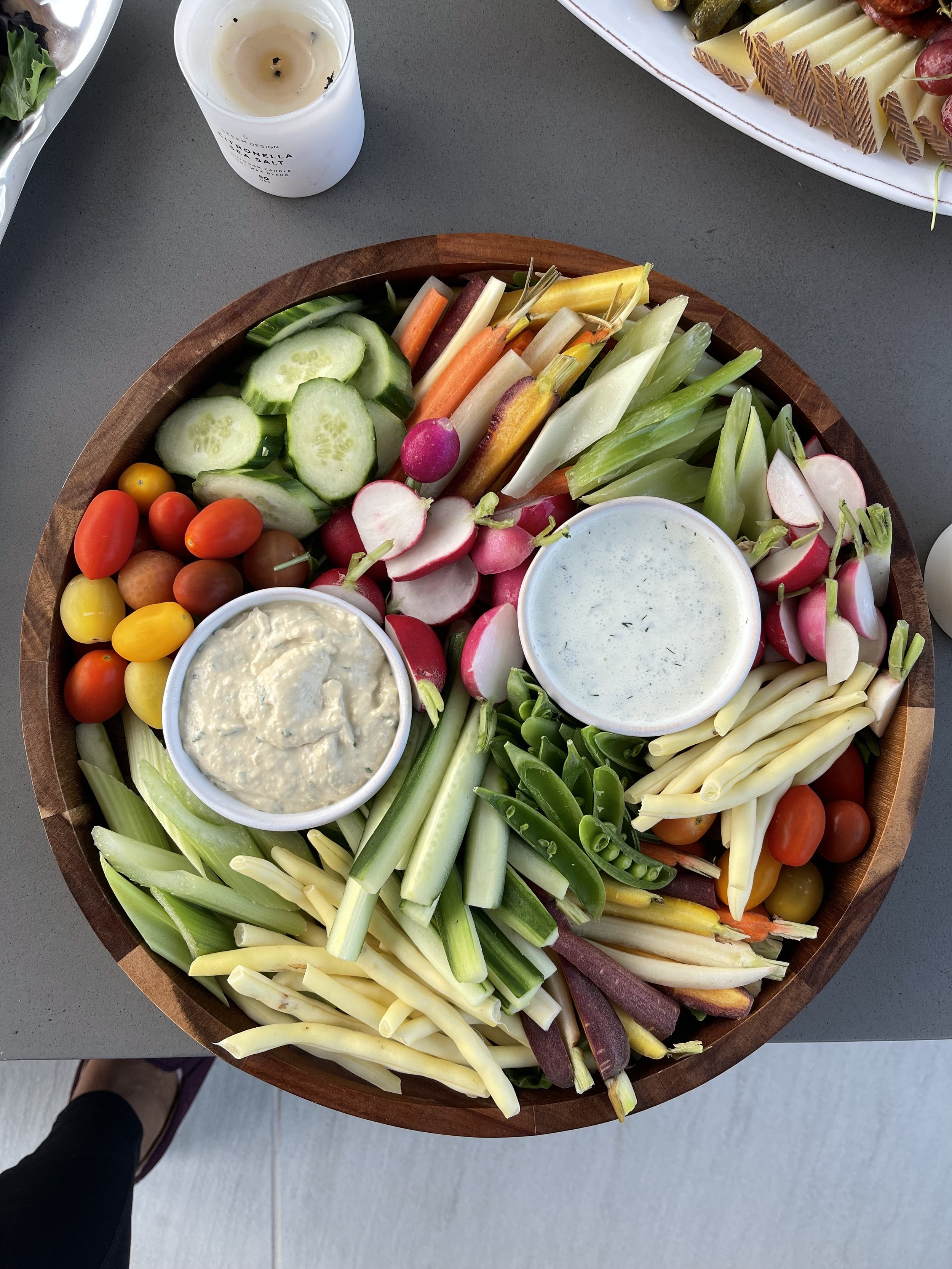 Wooden platter with a variety of fresh vegetables including cherry tomatoes, cucumber slices, radishes, celery, and colorful carrots, accompanied by two bowls of dip.