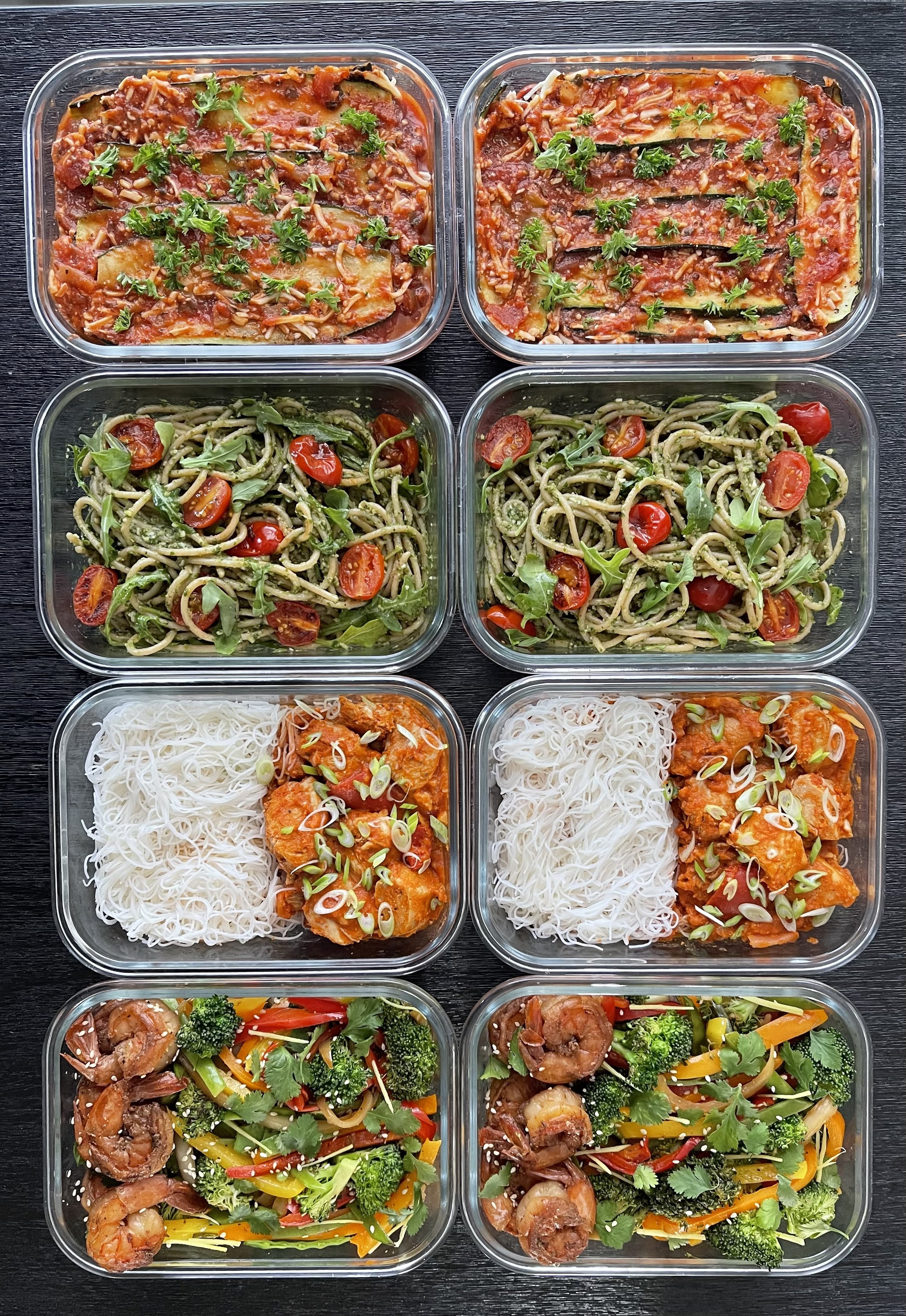 Meal prep with glass containers featuring lasagna, pesto pasta with cherry tomatoes and arugula, rice noodles with curry chicken, and shrimp with stir-fried vegetables.