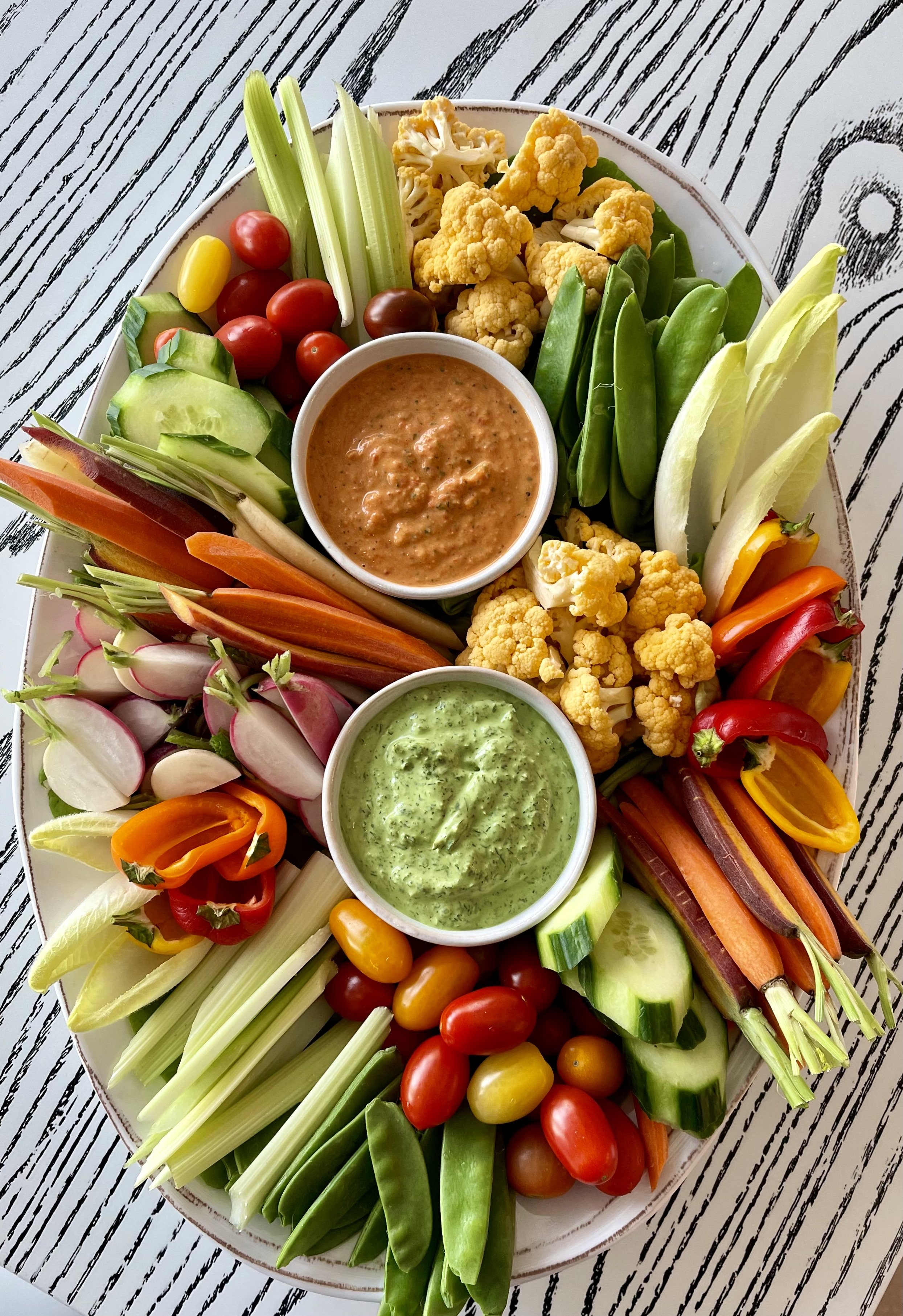 A platter of fresh vegetables including cucumbers, cherry tomatoes, celery, carrots, radishes, cauliflower, snap peas, and bell peppers. The platter features two bowls of dip, one red and one green.
