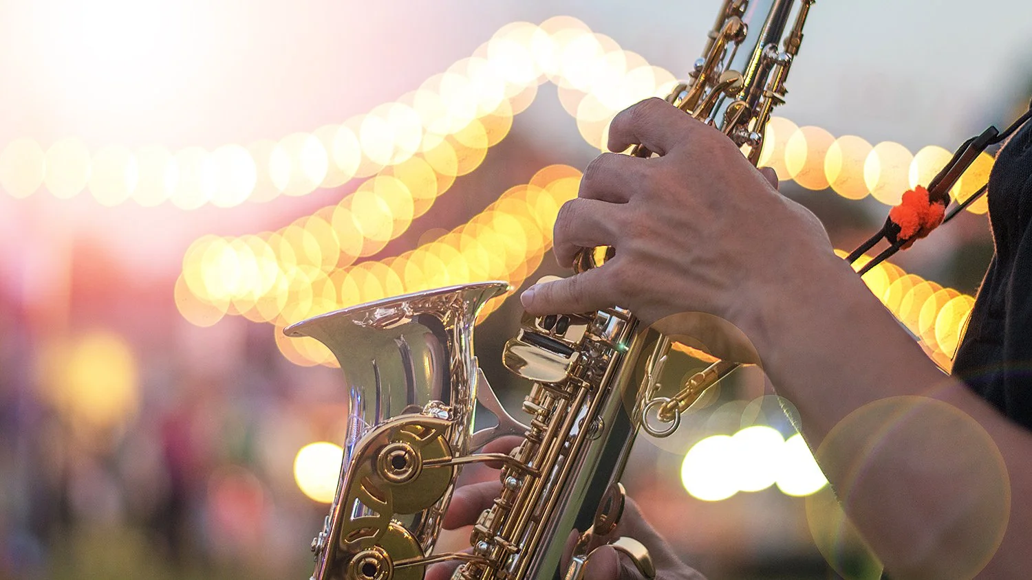 Close-up of a person playing a gold-colored saxophone outdoors during evening, with bokeh lights in the background.