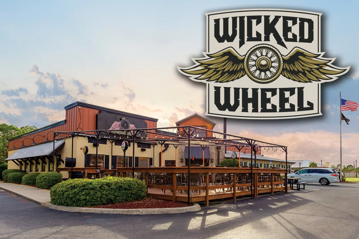 Outside of a restaurant called Wicked Wheel with a large sign, outdoor patio with seating, and an American flag in the background.