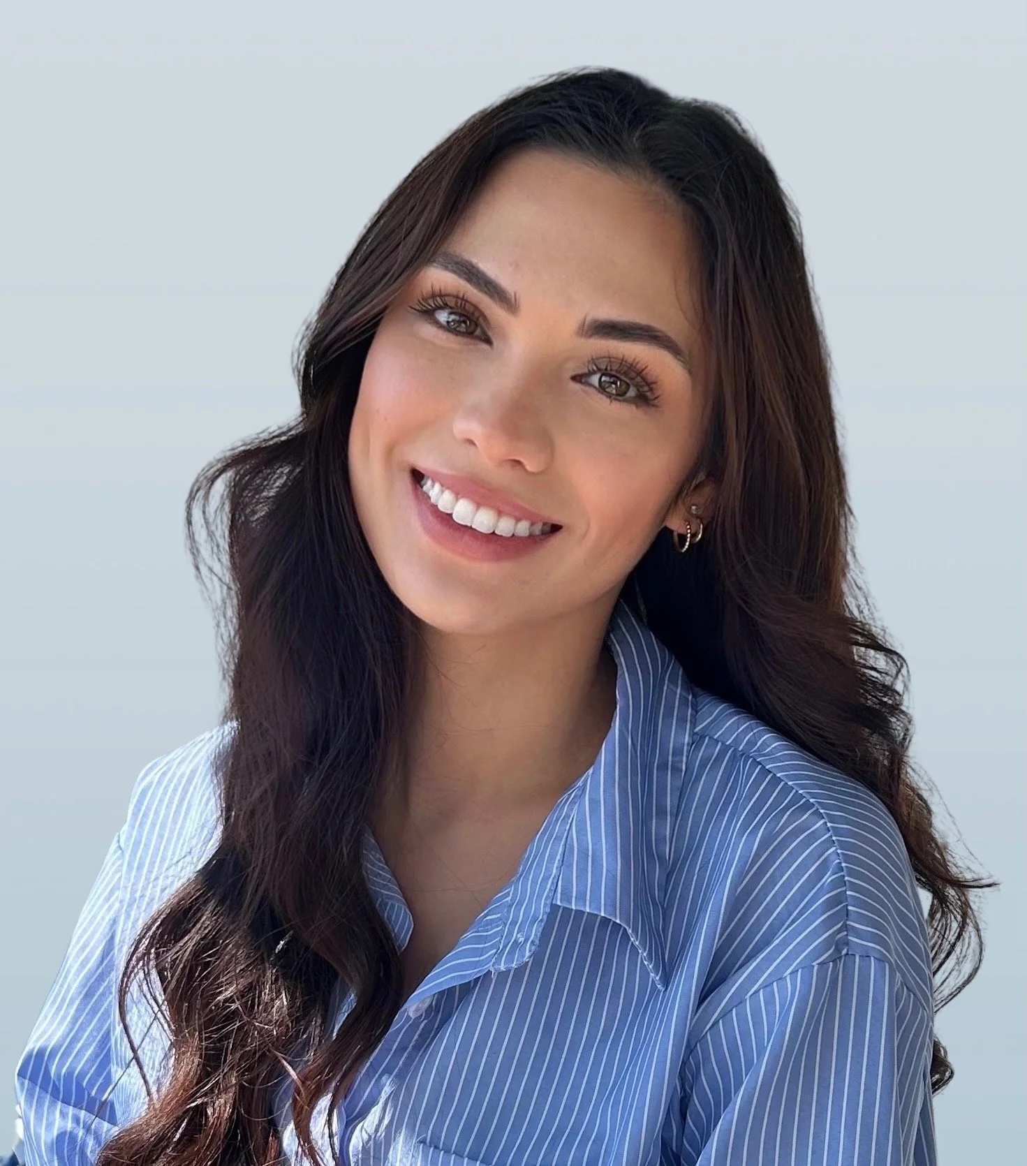 A smiling woman with long dark hair wearing a blue and white striped shirt, posing against a light gray background.