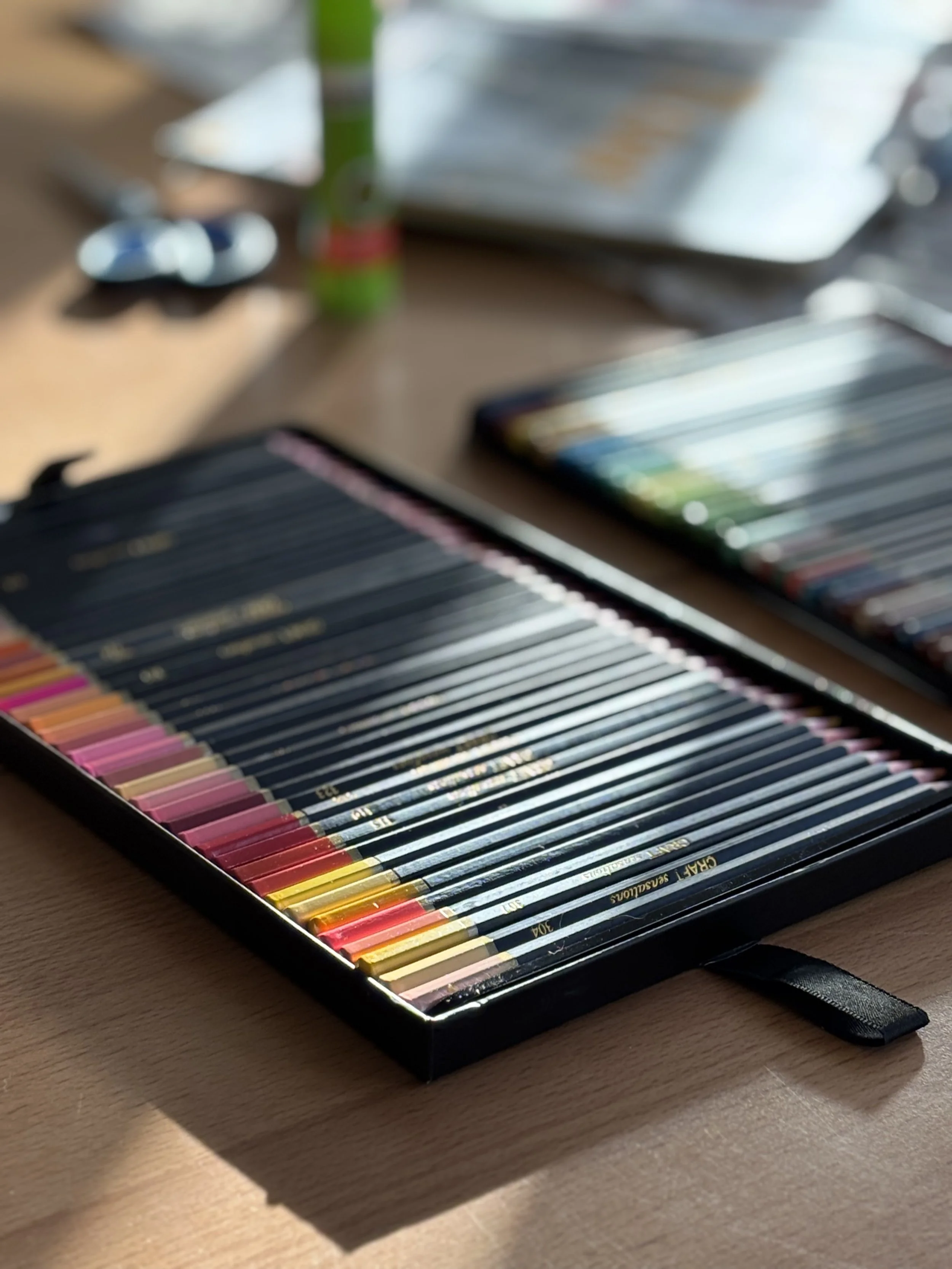 Een close-up van een set kleurpotloden in een zwarte case op een houten tafel, met andere ksdes en een laptop op de achtergrond, onder natuurlijk licht.