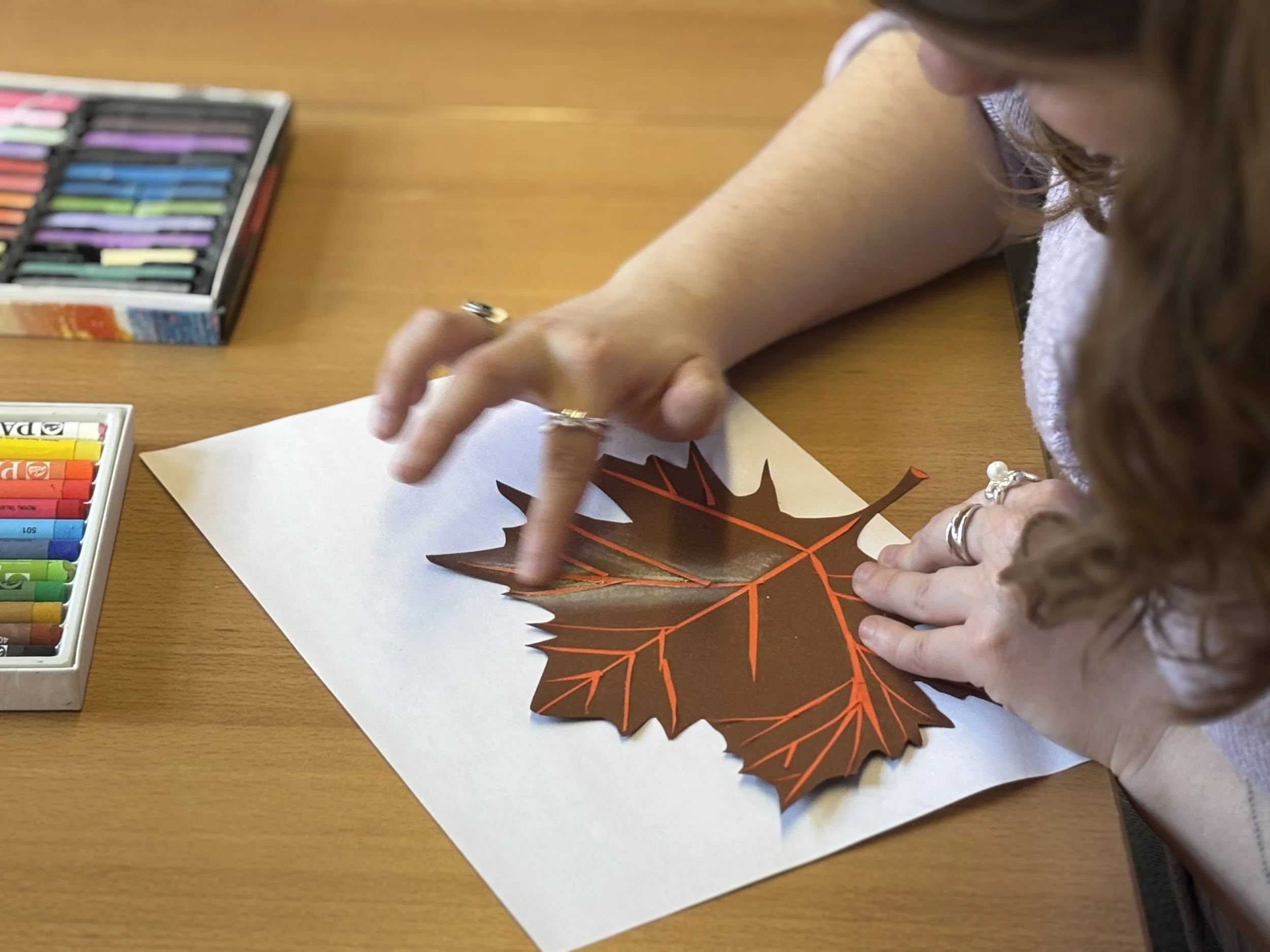 Een persoon maakt een kunstwerk van een herfstblad met oranje stiften op een wit stuk papier. Ernaast liggen uiteenlopende kleurkrijtjes en pastelkrijten op een houten tafel.