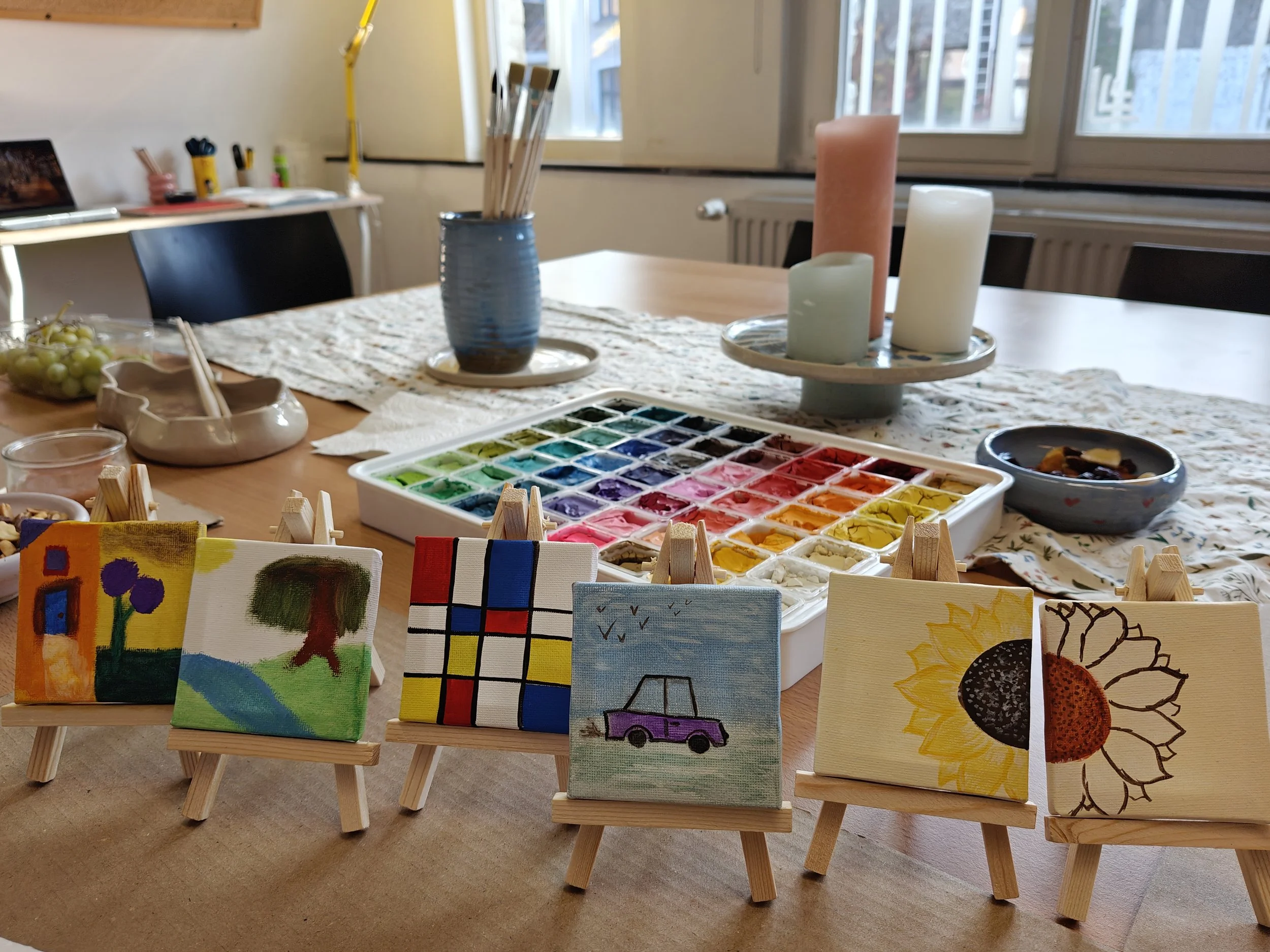 Kleine schilderijen op miniatuur schildereasjes met diverse abstracte en figuratieve kunst, geplaatst op een tafel met waterverf en schildermaterialen.