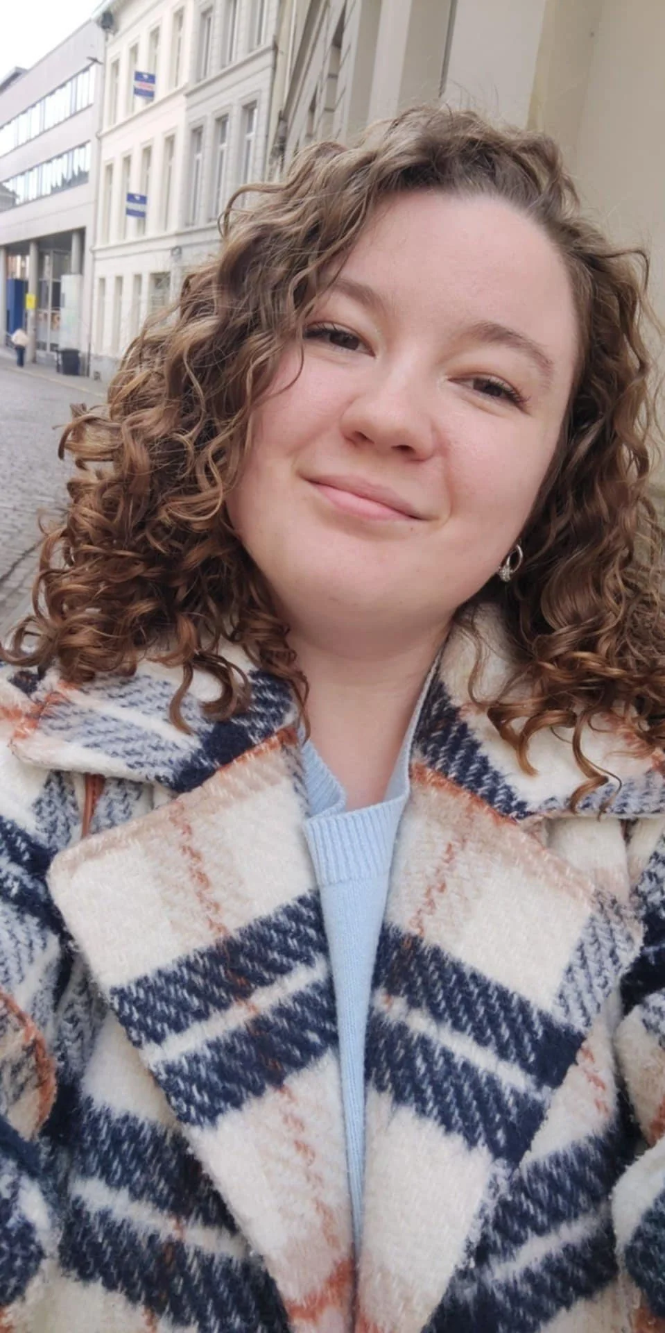 Een meisje met grotendeels gekrulde bruin haar, lachend, buiten op een straat met gebouwen op de achtergrond, draagt een schaakbordpatroon jas.