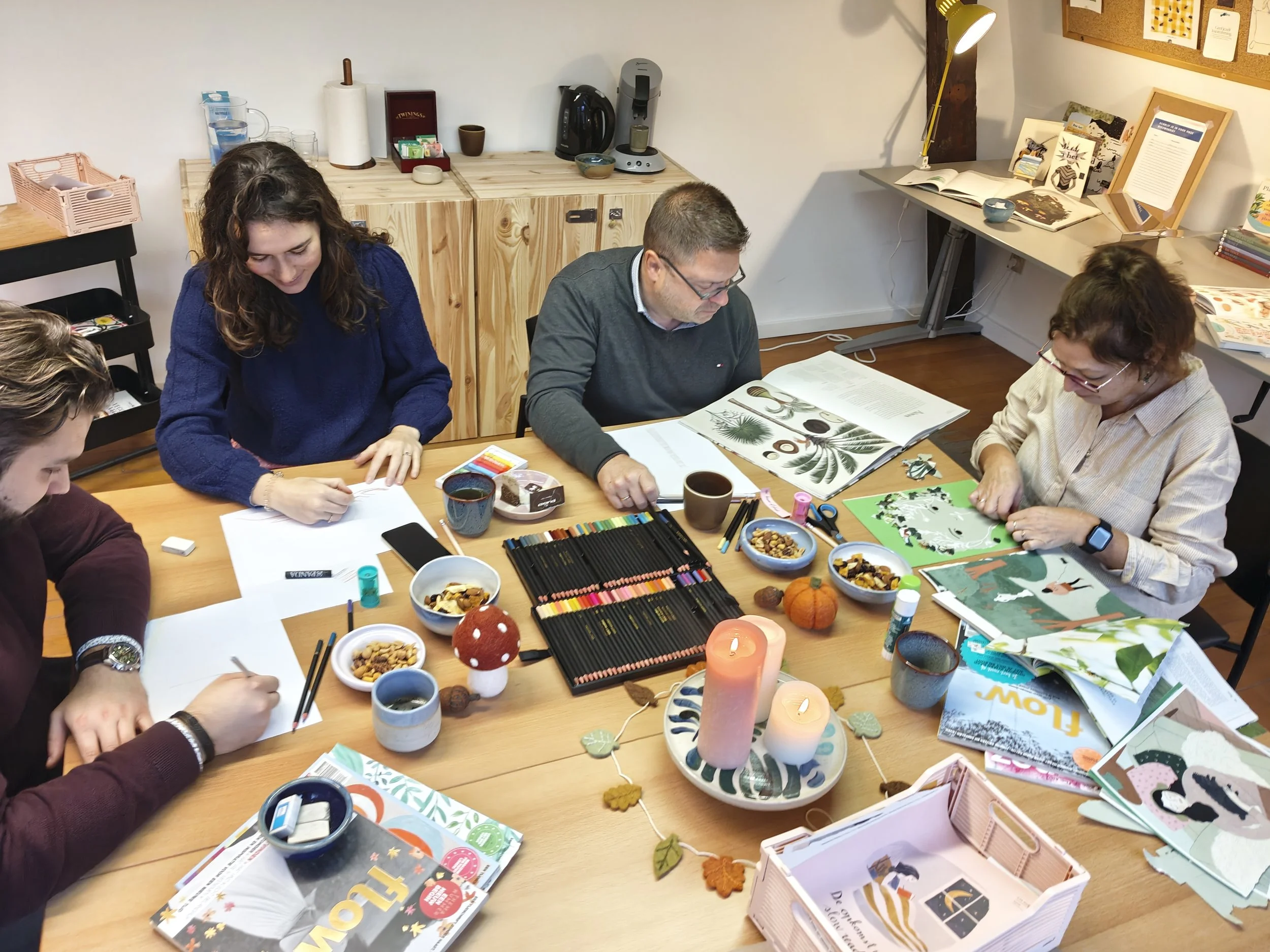 Vijf mensen zitten aan een tafel die versierd is met kaarsen en herfstdecoraties, terwijl ze tekenen en knutselen met verschillende materialen en boeken, in een gezellige en creatieve groepsactiviteit.
