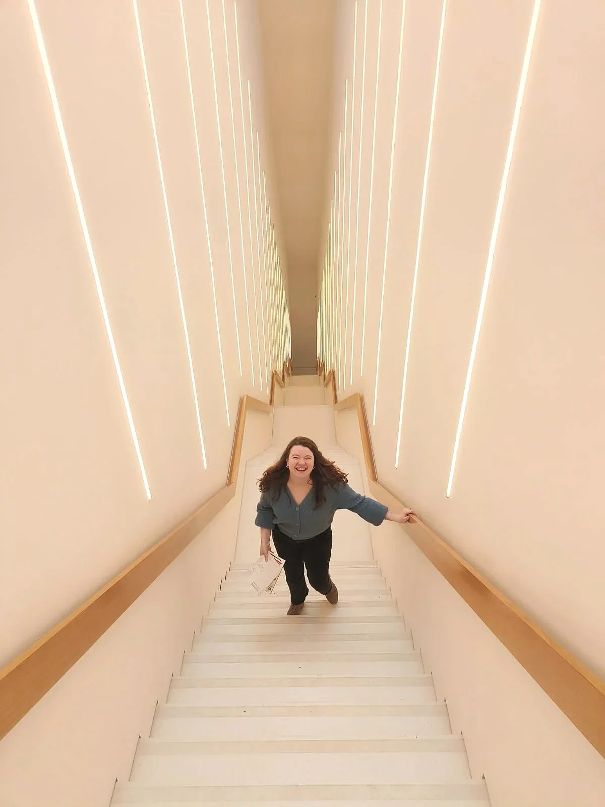 Vrouw die de trap op loopt in een verlichte, moderne traphal met verticale lichtlijnen op de muur in een museum
