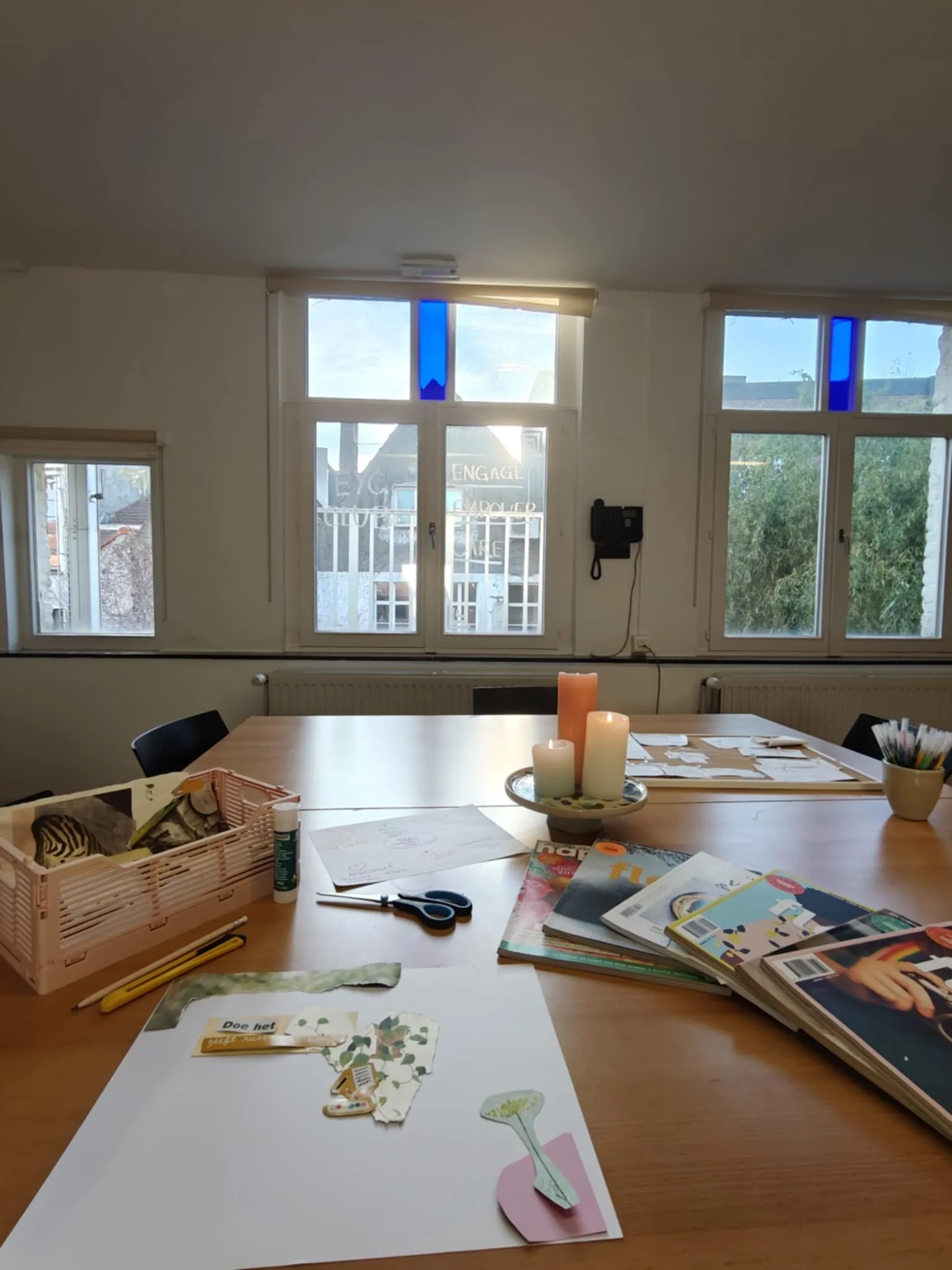 Binnenkant van het atelier met een houten tafel met knutselspullen, kaarsen en tijdschriften, met ramen en zonlicht valt mooi naar binnen.