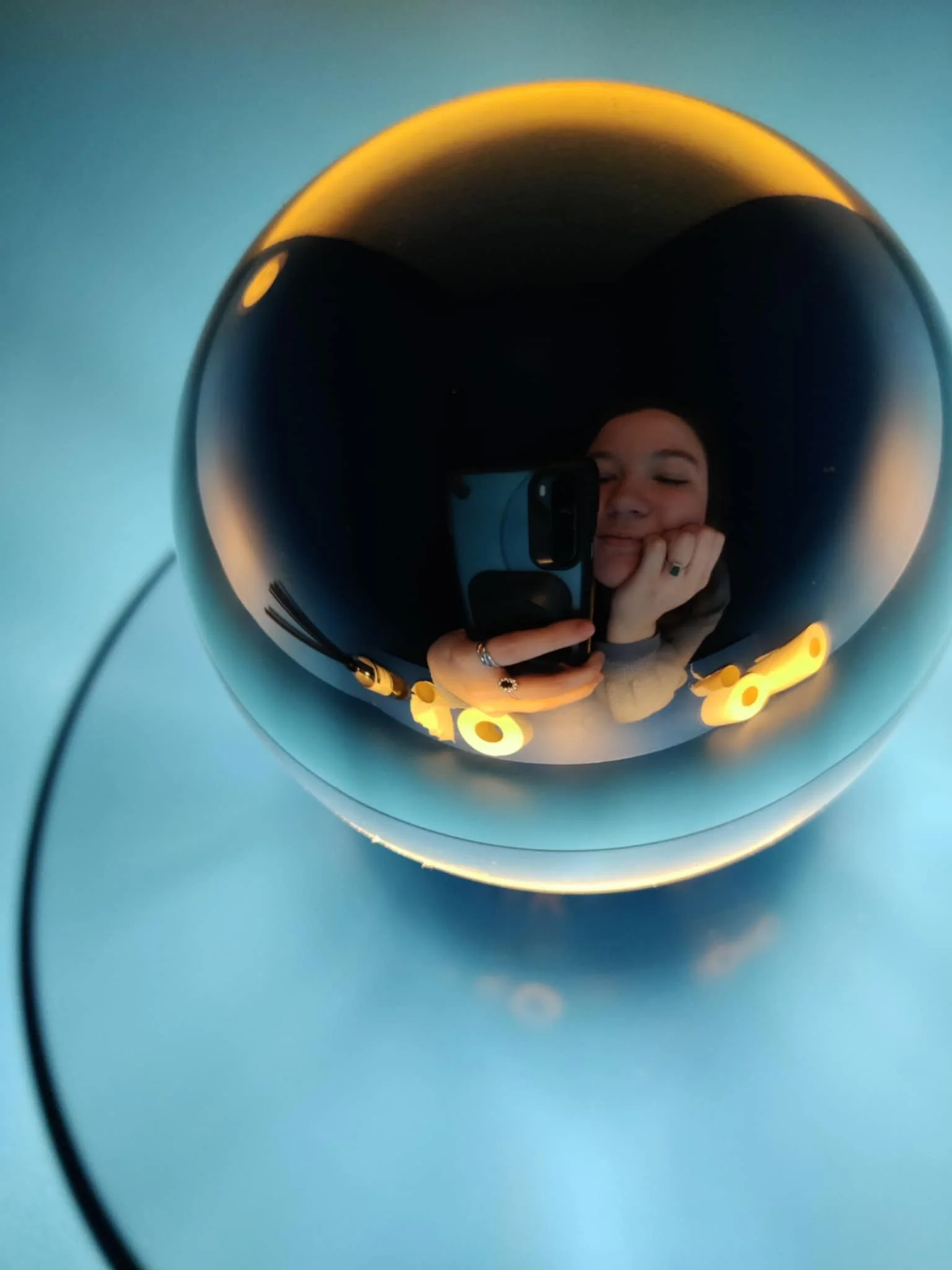 Eline zichtbaar via een spiegel in een modern, zwart, ronde lamp met gele details, met een blauwe achtergrond.