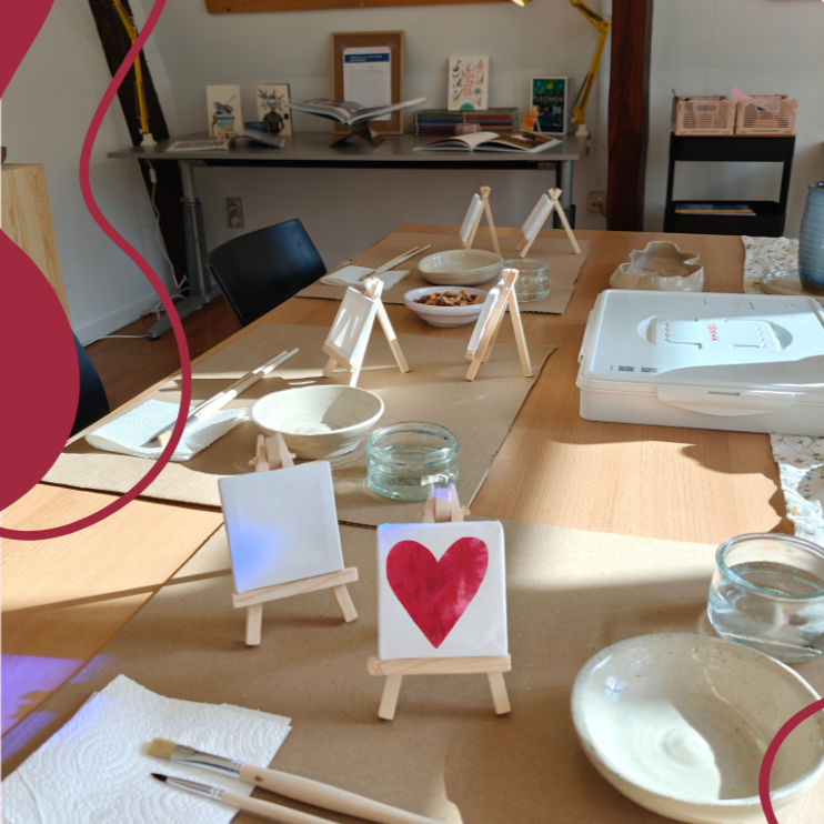 Een tafel met mini schilderdoeken, kleine ezels en schildermaterialen, klaar voor een schilderworkshop of creatieve sessie. Er staat een hartje op geschilderd.