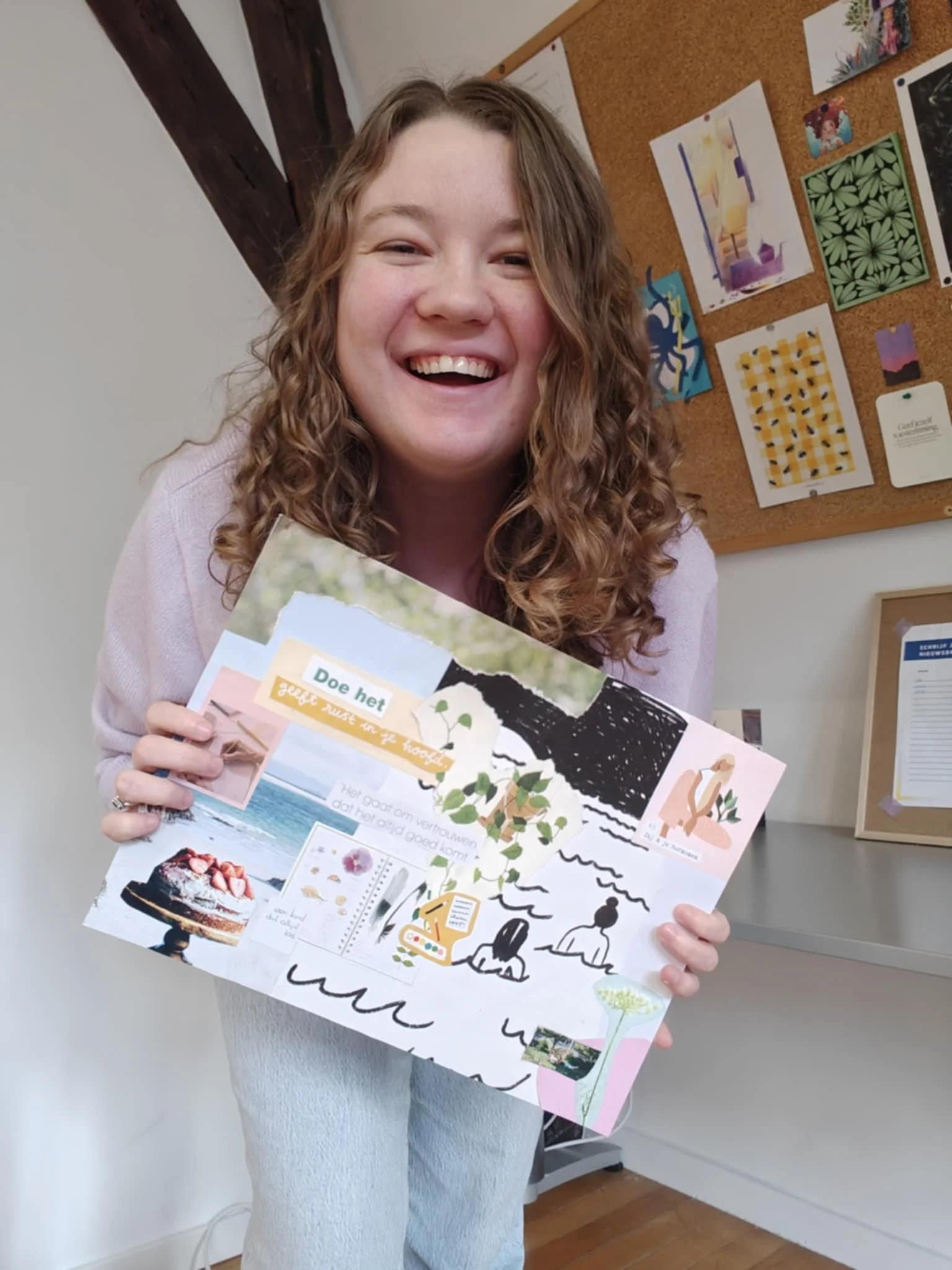 Vrolijke jonge vrouw met lange, gekrulde haar die vrolijk een collage van foto's en tekeningen vasthoudt in een kamer met een prikbord en speelkaarten op de achtergrond.