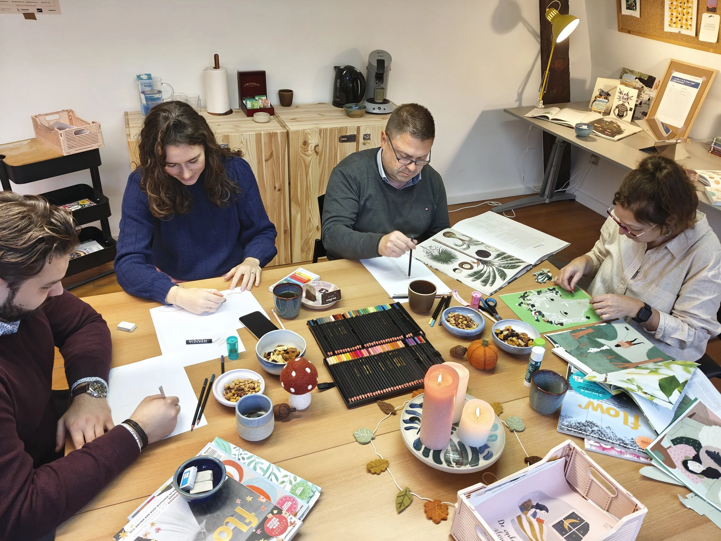 Vijf mensen zitten aan een tafel die versierd is met kaarsen en herfstdecoraties, terwijl ze tekenen en knutselen met verschillende materialen en boeken, in een gezellige en creatieve groepsactiviteit.