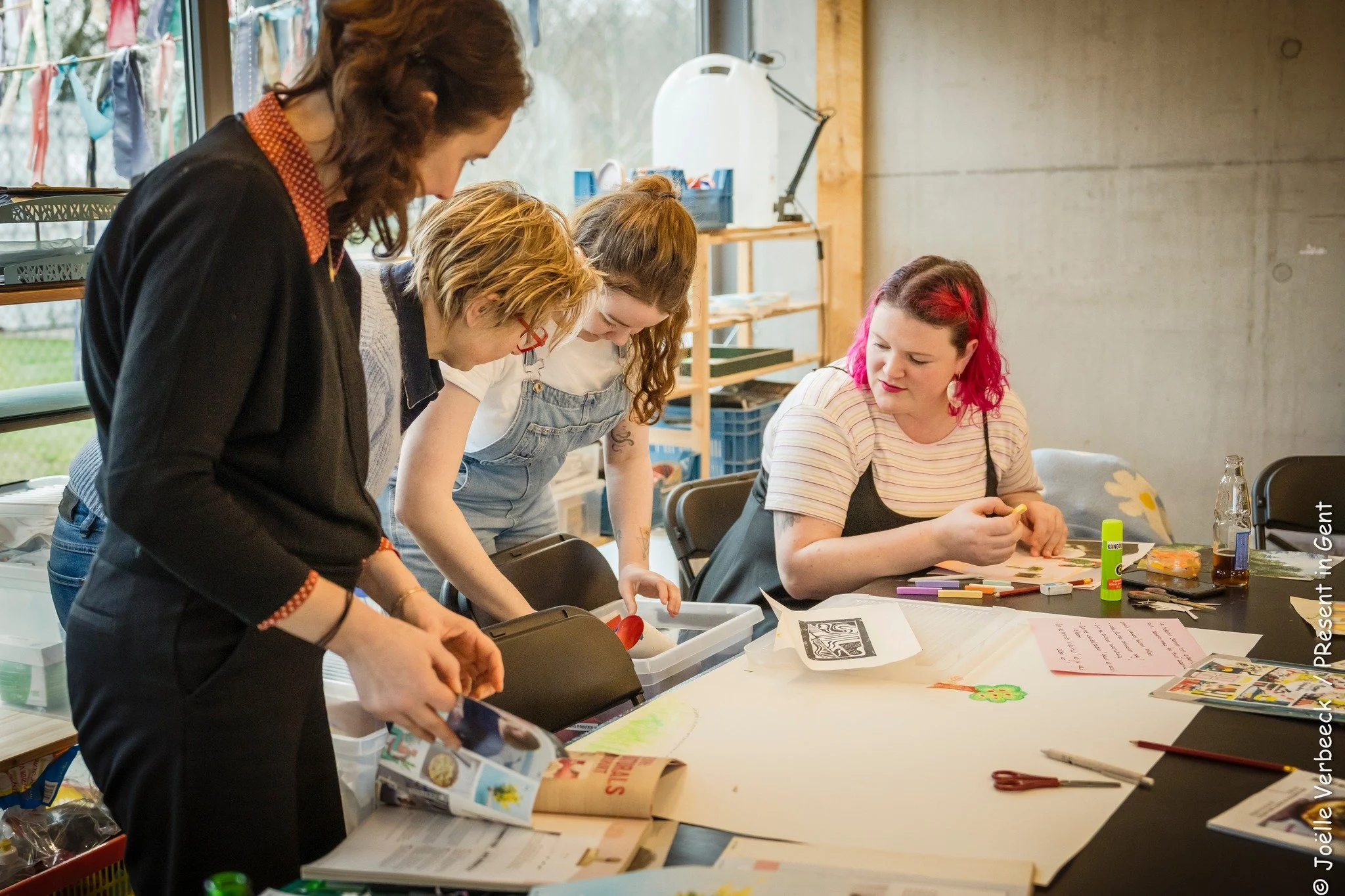 Vijf mensen werken samen aan een creatieve knutselproject aan een tafel met knutselmaterialen, papieren en tijdschriften in een indoors ruimte.