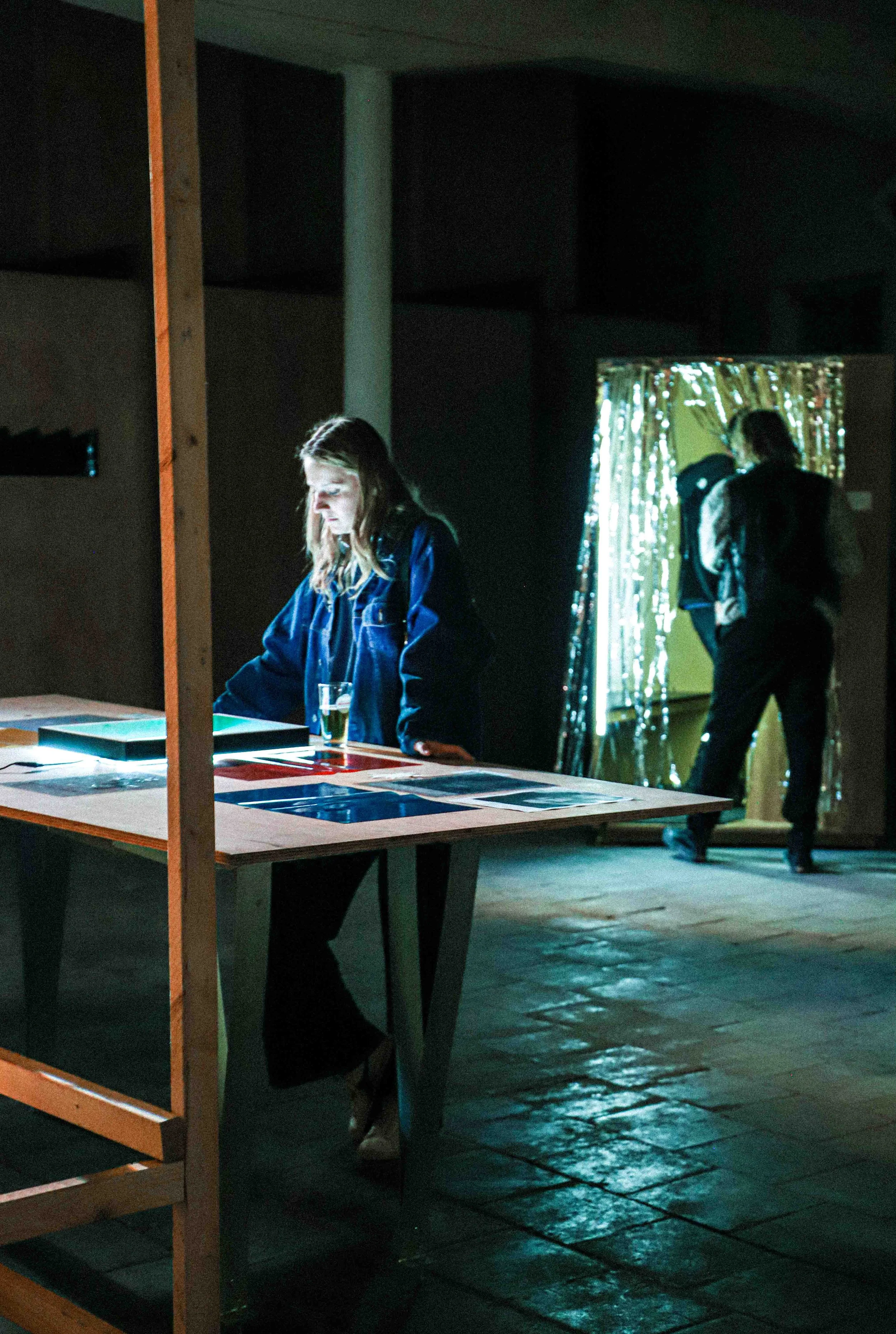 Een vrouw die naar een tafel waar kunst op ligt, terwijl op de achtergrond mensen voorbij lopen in een donkere ruimte met een decoratief scherm met verlichting.