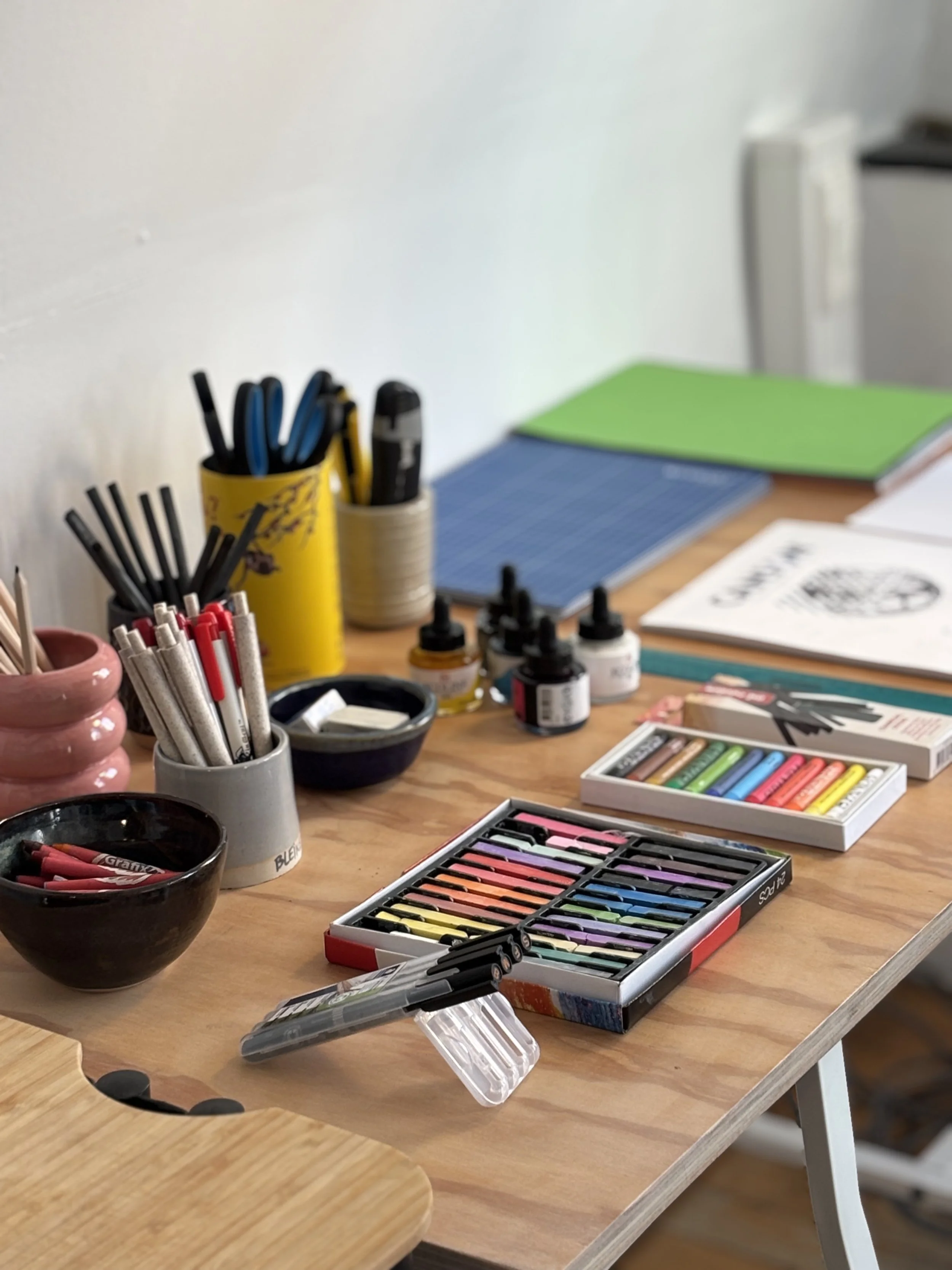 Kunstartikelen en pastel krijtjes op een houten tafel, inclusief potloden, verf, verfborstels, en papier.