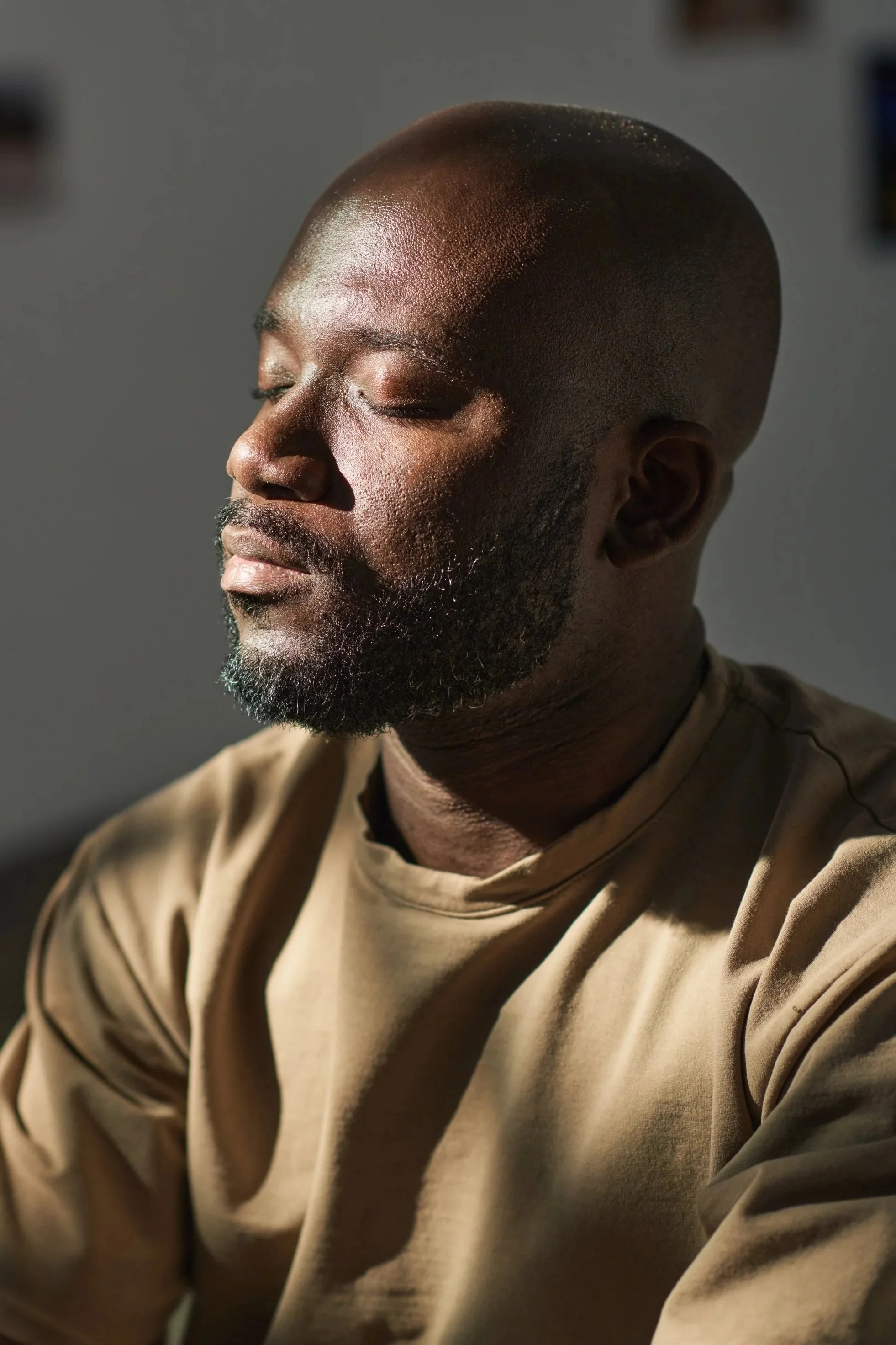 A close-up side profile of a man with closed eyes, wearing a beige shirt and having a shaved head and beard, with a soft light illuminating his face.