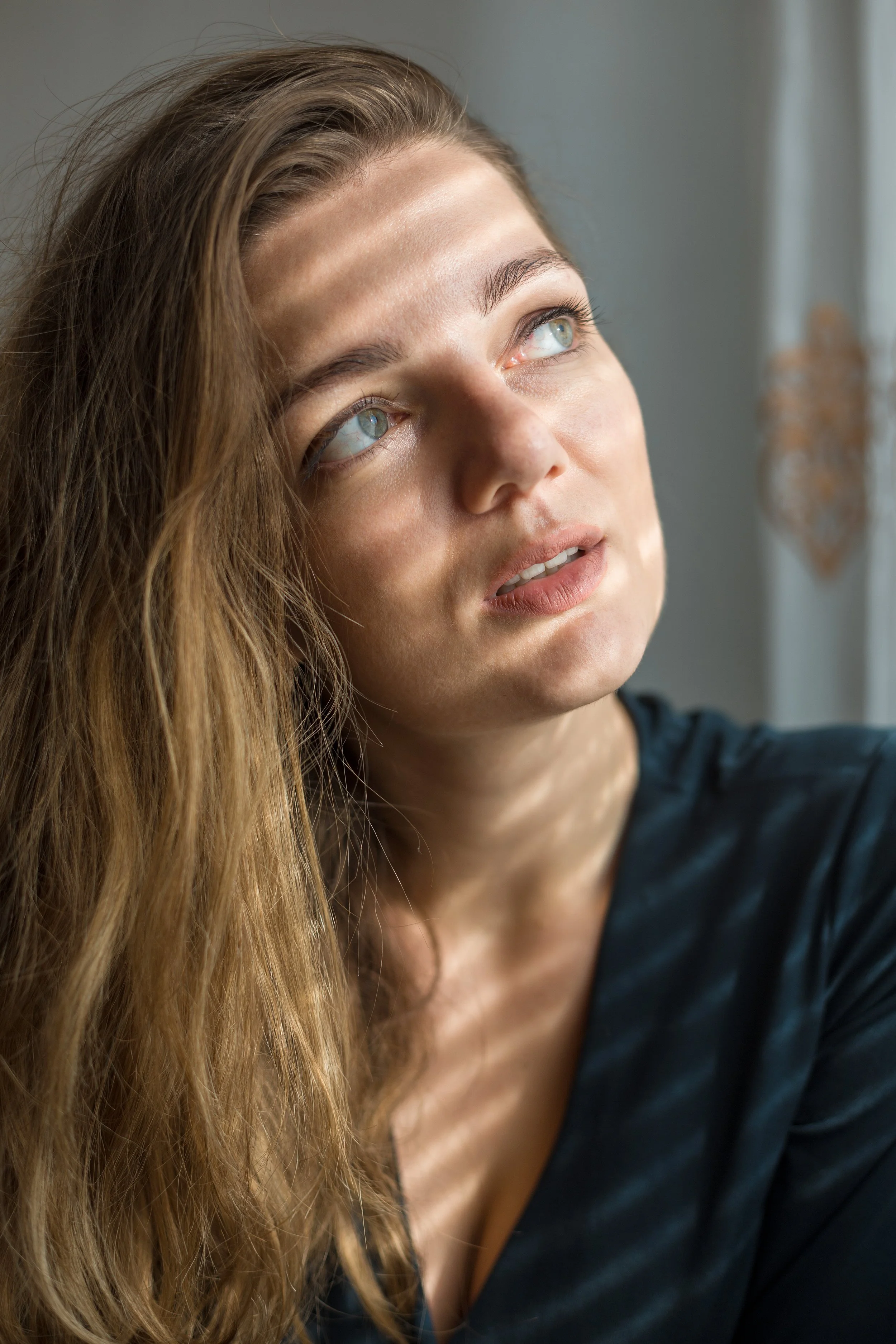 Close-up of a young woman with long wavy blonde hair, light skin, looking upwards and to the left, with bright blue eyes, wearing a dark shirt, standing near a window with light-filtered background.