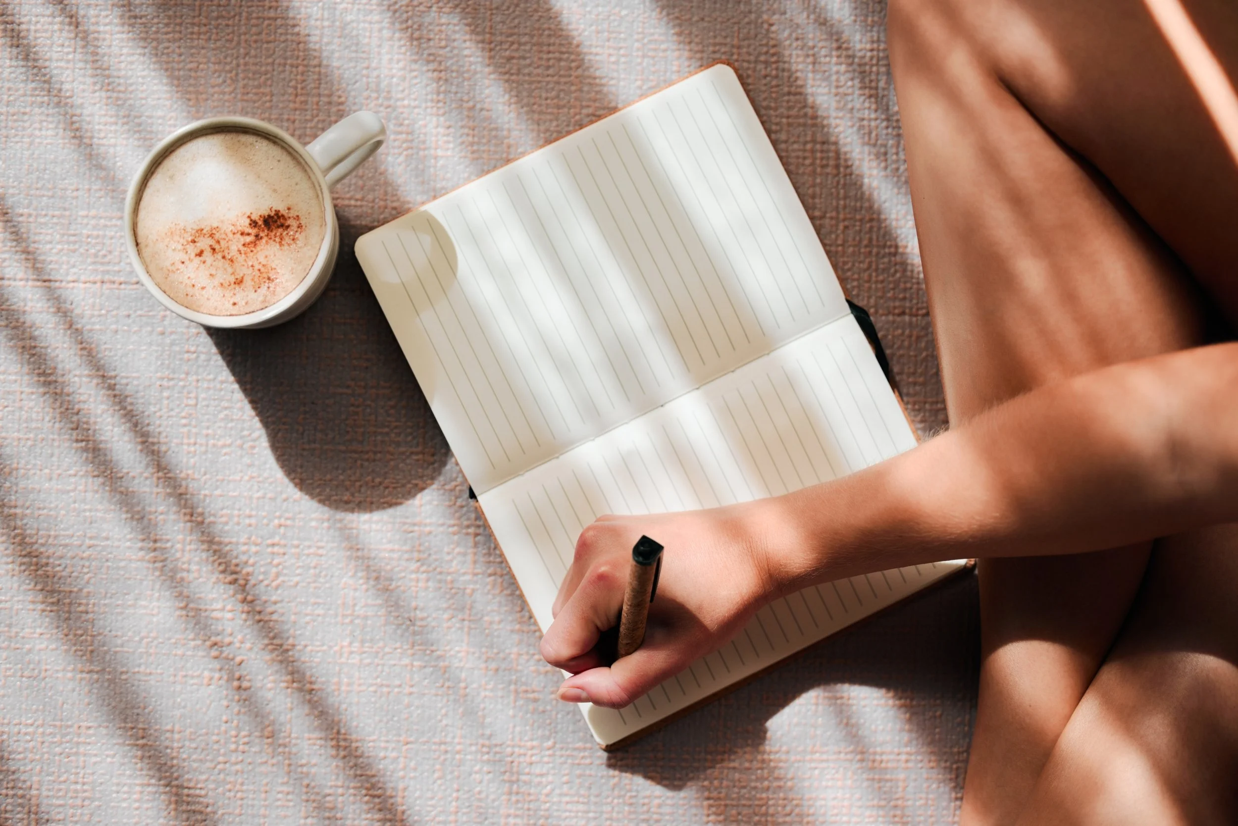 A person sitting on a textured pink blanket, writing in an open lined notebook with a black pen. There is a white mug filled with a frothy beverage, topped with red spice, placed next to the notebook.