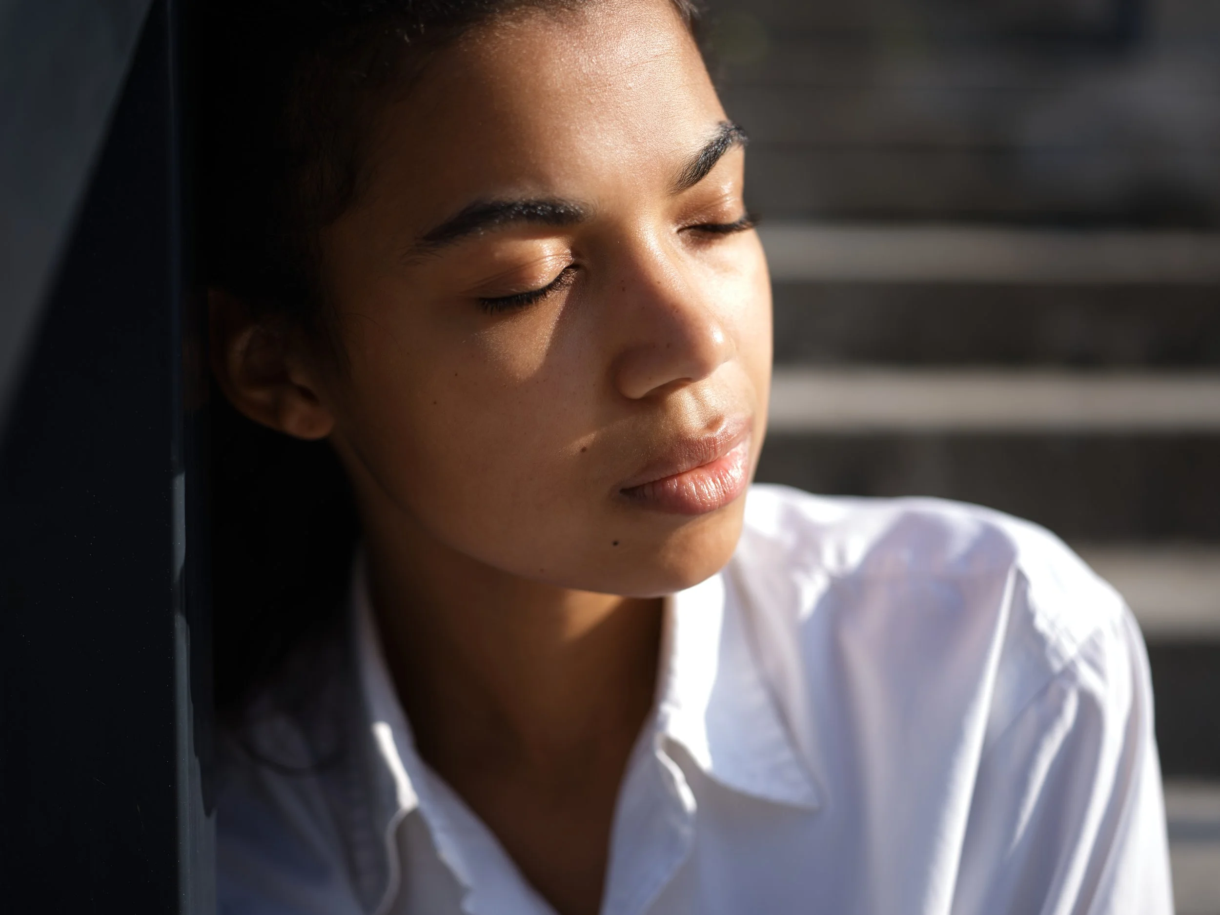 Close-up of a woman with medium dark skin, closed eyes, and dark hair, wearing a white shirt, with sunlight highlighting her face and a blurred background.