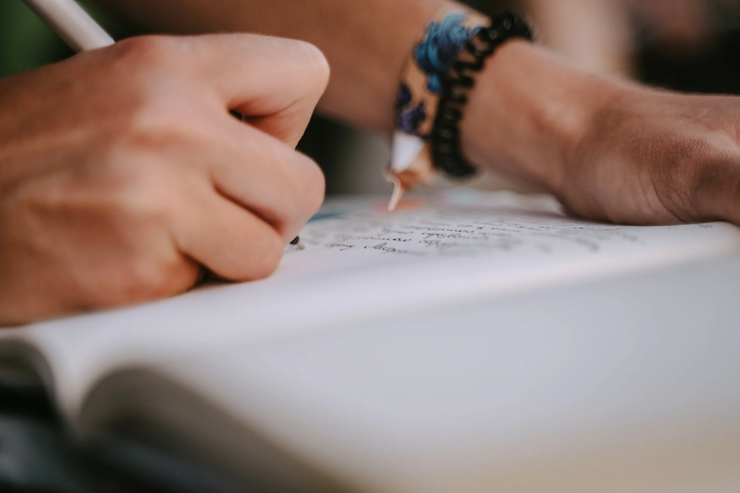 Close-up of a person writing on paper with a pen, wearing a black beaded bracelet.
