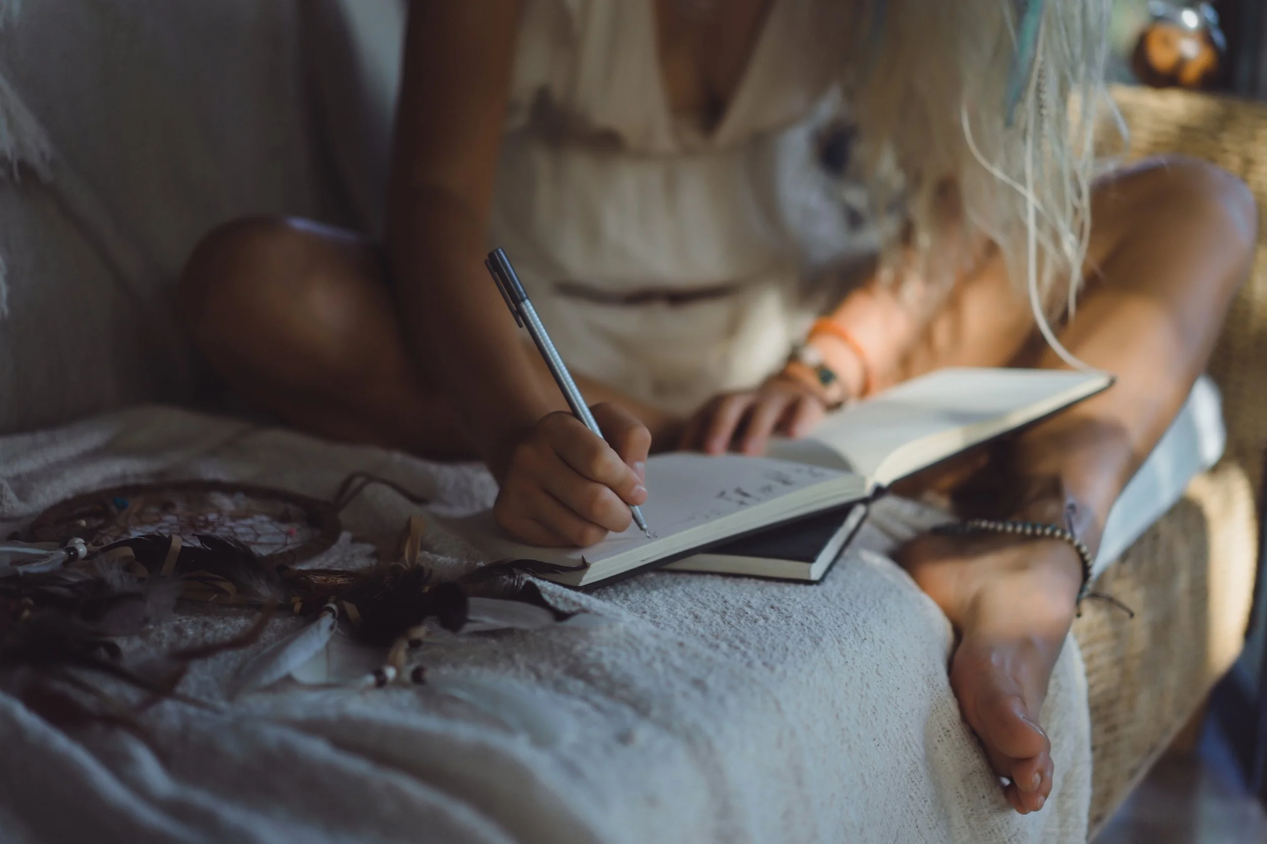 A person sitting on a blanket appears to be writing in a notebook, with long hair and wearing a bracelet, surrounded by other belongings, with a sunlit outdoor setting.