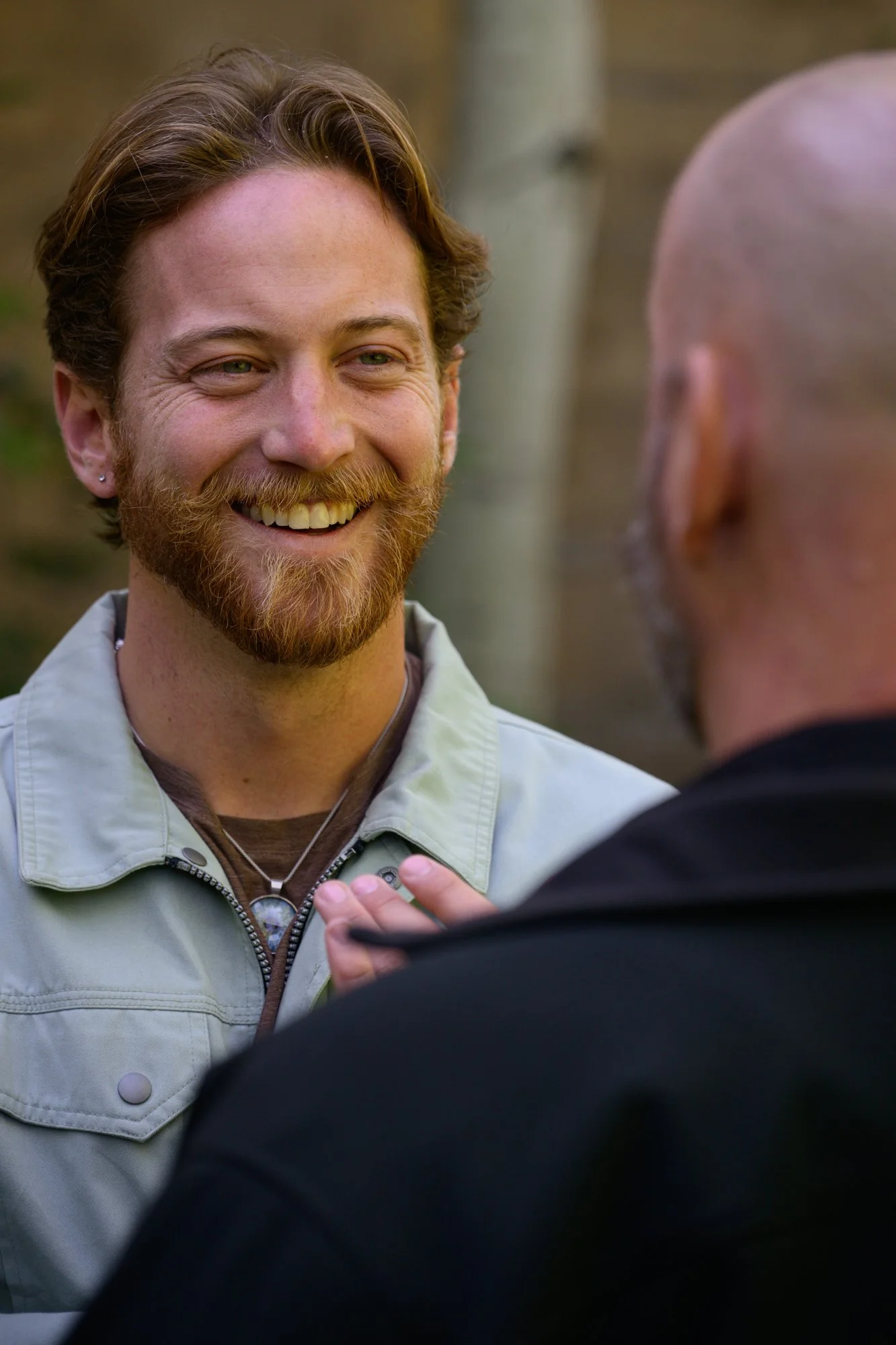 Two men facing each other outdoors, one smiling, engaged in conversation.