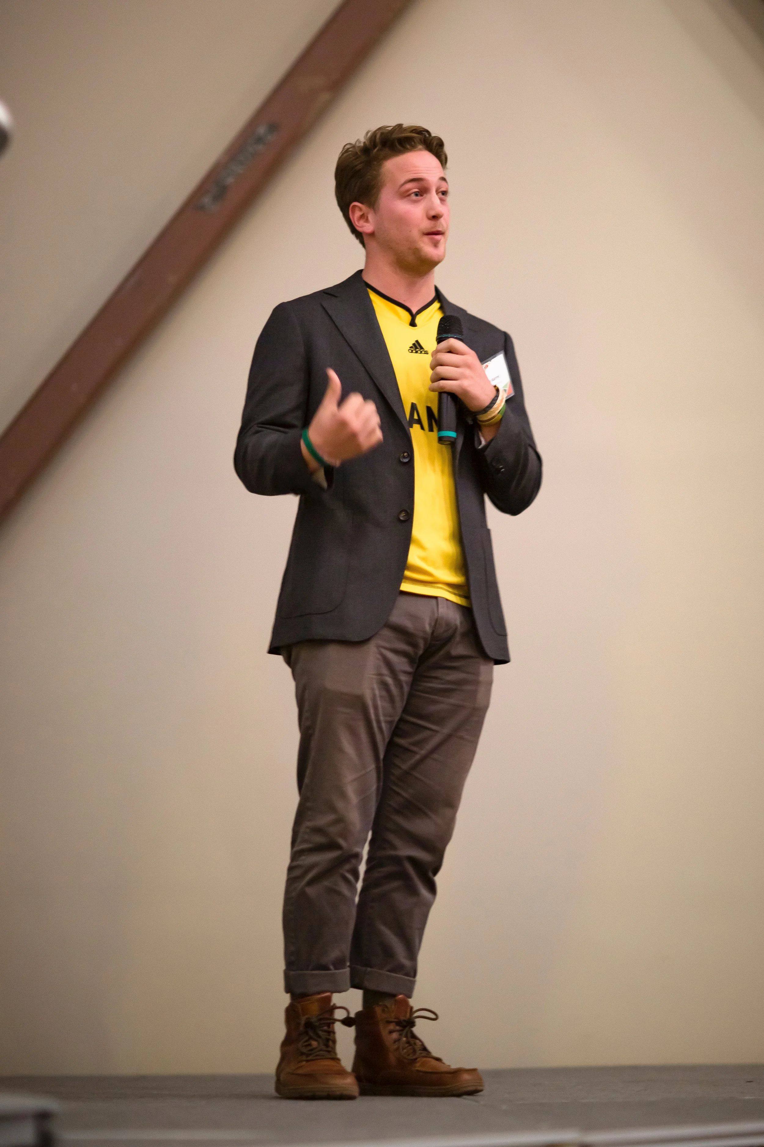 Tom Karrel, wearing a yellow Adidas soccer jersey, a dark blazer, brown pants, and tan boots, standing on a stage with a beige wall background, holding a microphone in his right hand, and gesturing with his left hand.