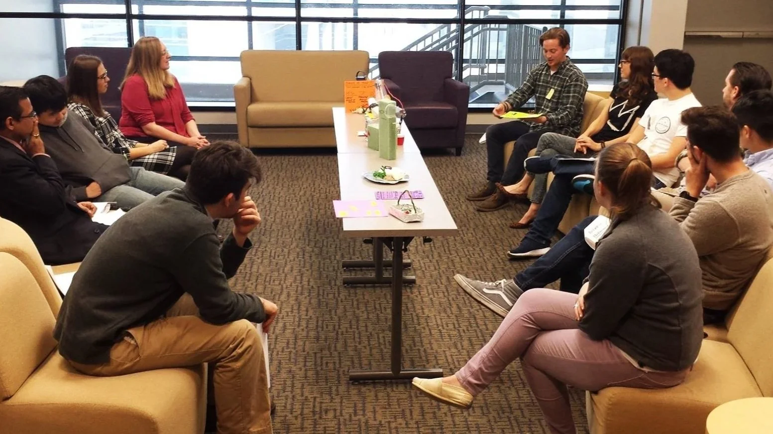 A group of people sitting in a semi-circle around a long table, engaged in a discussion or presentation in a modern office or conference room with large windows.
