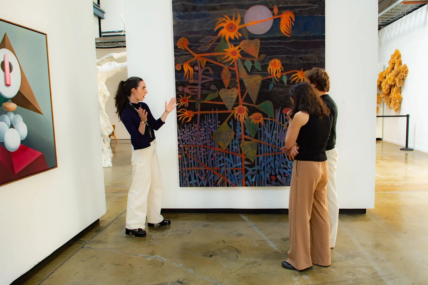 Une guide touristique explique une peinture abstraite à un groupe de trois visiteurs dans une galerie d'art, visite et médiation, visite de groupe et scolaire