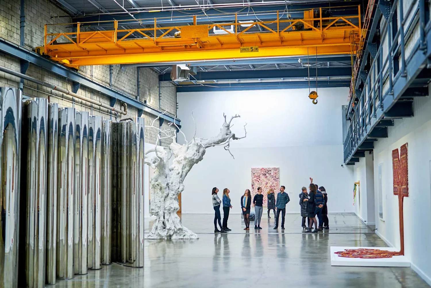 Un groupe de personnes dans une galerie d'art moderne avec une sculpture d'arbre blanc, des œuvres d'art colorées, un plafond industriel, et un pont roulant orange.