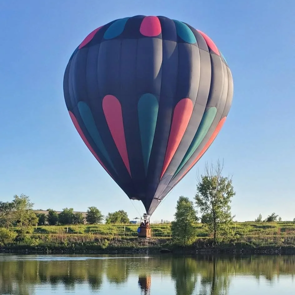A hot air balloon with pink, teal, and navy vertical stripes flying in the sky above trees.