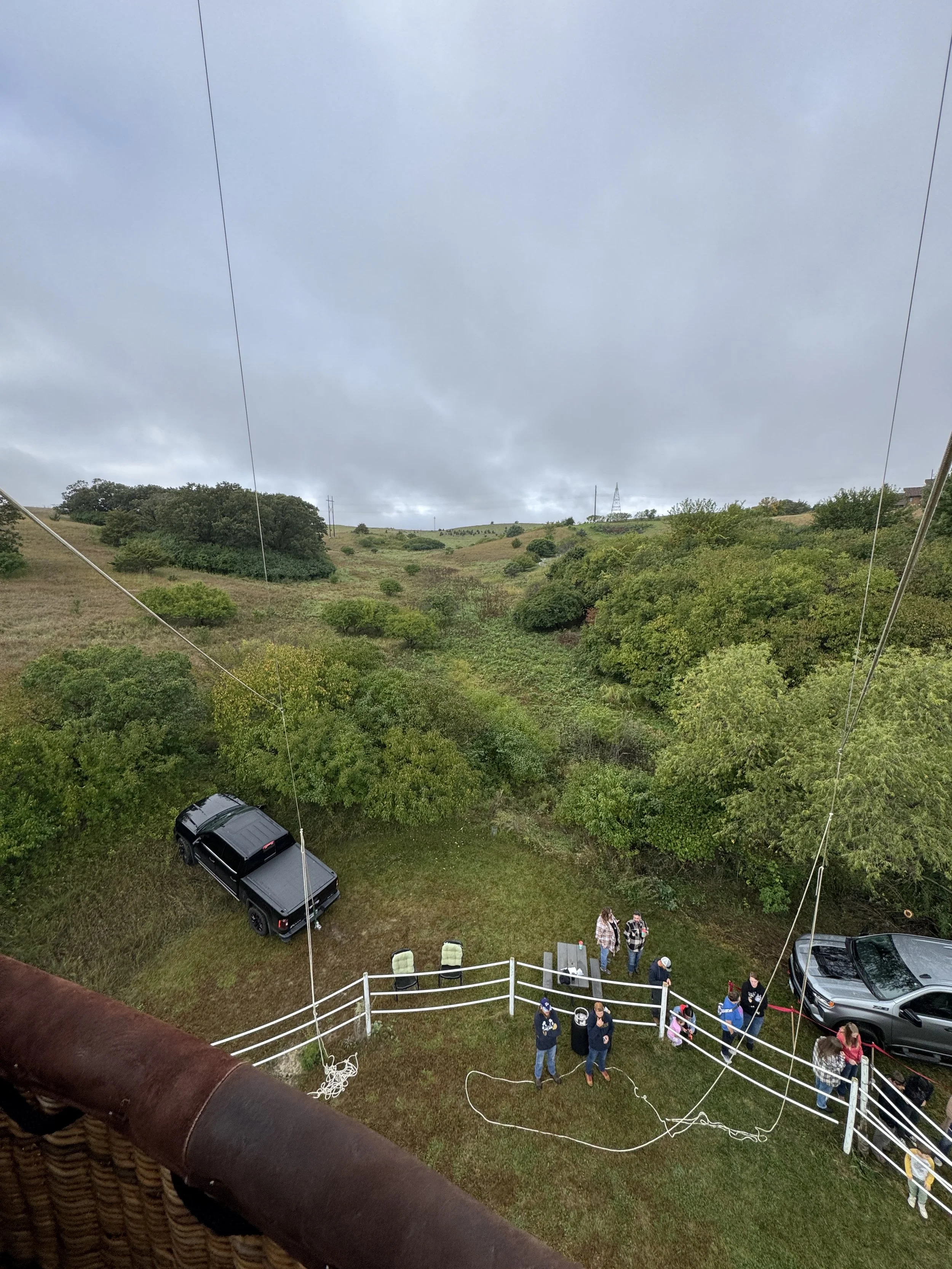 A view from a hot air balloon looking down at a group of people on the grass near parked cars, with a lush green landscape and cloudy sky in the background.