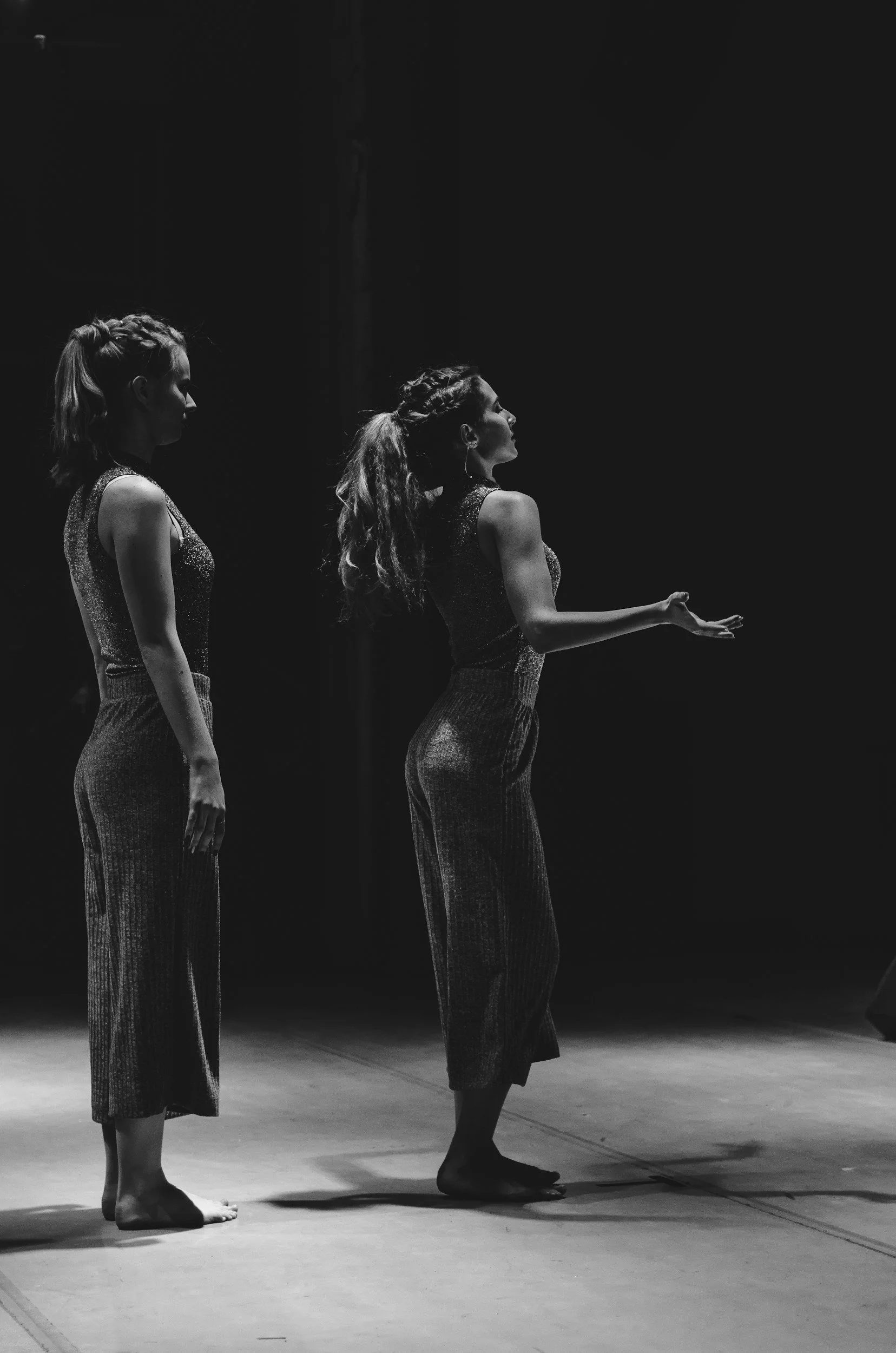 A woman leading a yoga or meditation class in a bright, urban studio with large windows, seated on a purple mat, demonstrating a hand gesture to the participants.