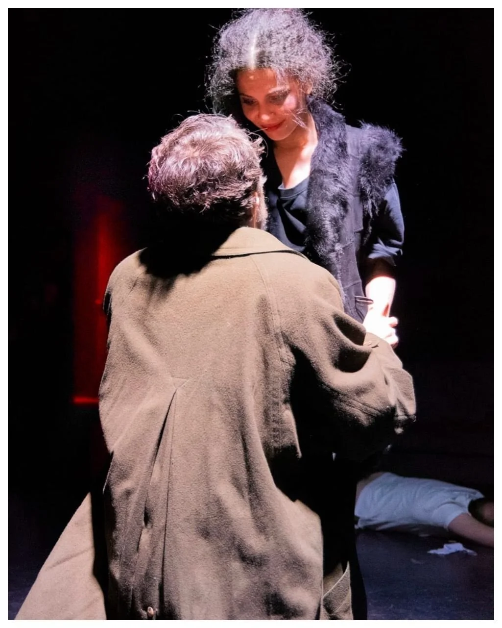 A woman leading a yoga or meditation class in a bright, urban studio with large windows, seated on a purple mat, demonstrating a hand gesture to the participants.