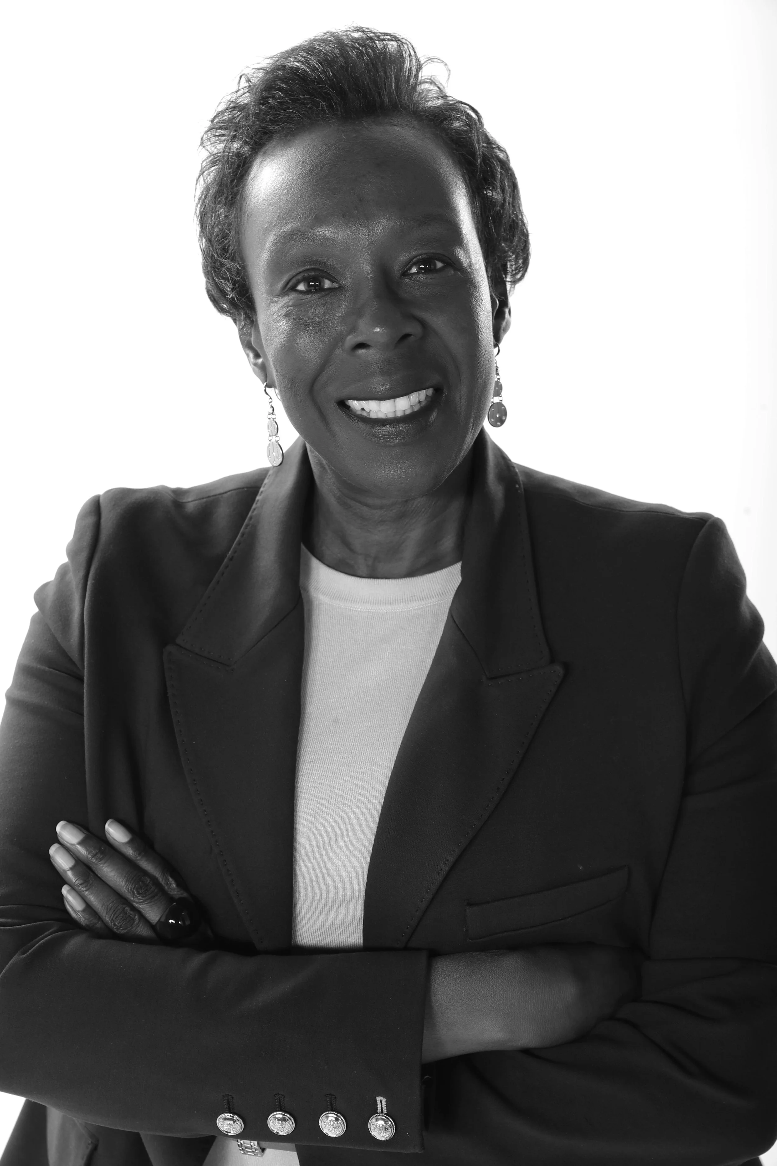 A woman in a beige blazer with a yellow scarf, speaking and gesturing with her hand in a meeting or presentation, with a man visible in the foreground.