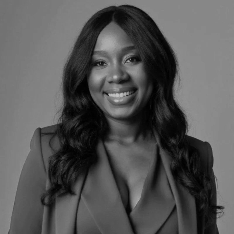 A woman leading a yoga or meditation class in a bright, urban studio with large windows, seated on a purple mat, demonstrating a hand gesture to the participants.