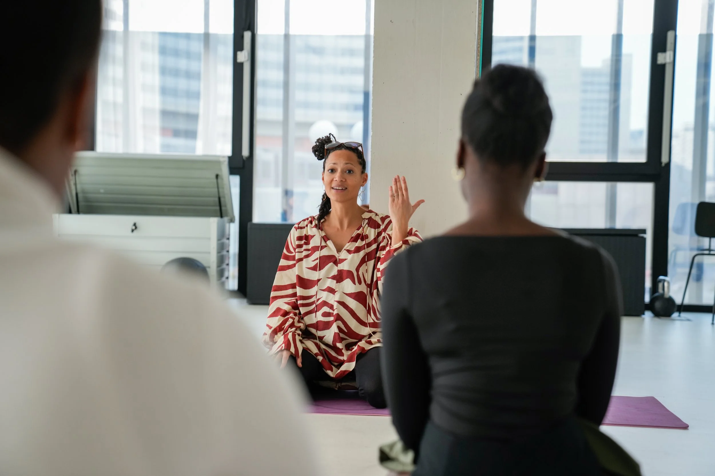 A woman in a patterned shirt sitting on a yoga mat and speaking to two people in a room with large windows.