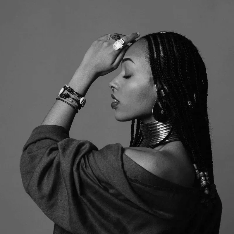 Two women practicing yoga inside. One woman is sitting cross-legged on a yoga mat, eyes closed, and singing or chanting, while the other is seated nearby, watching or listening. The setting is bright and airy with large windows and a view of an outdoor patio.
