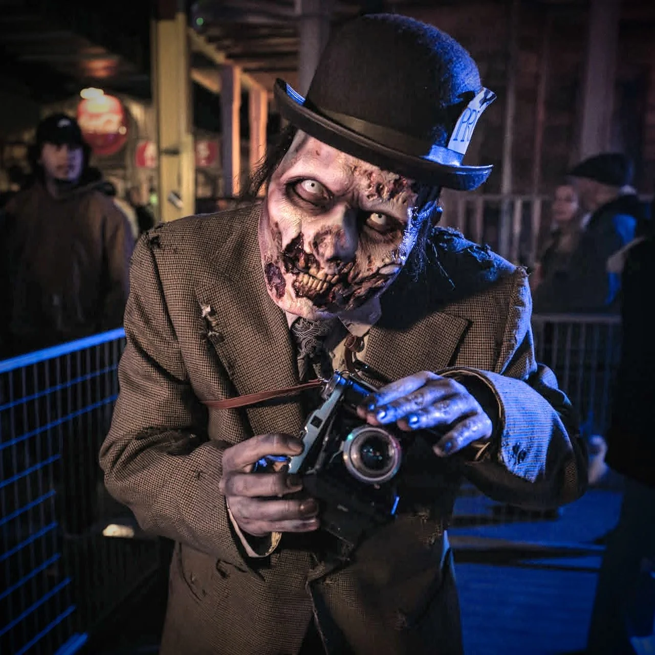 A person in zombie makeup with a skeletal face, wearing a brown jacket and a black top hat, holding a vintage camera. The background shows a dimly lit setting with a few people, possibly at a Halloween or themed event.