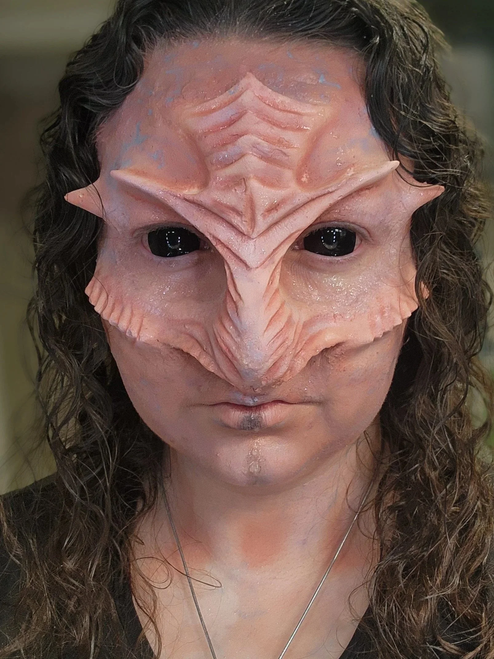 A person wearing a detailed aquatic makeup face prostheticwith large, black eyes, textured skin, and ridges on the forehead and around the face, with long, curly hair.