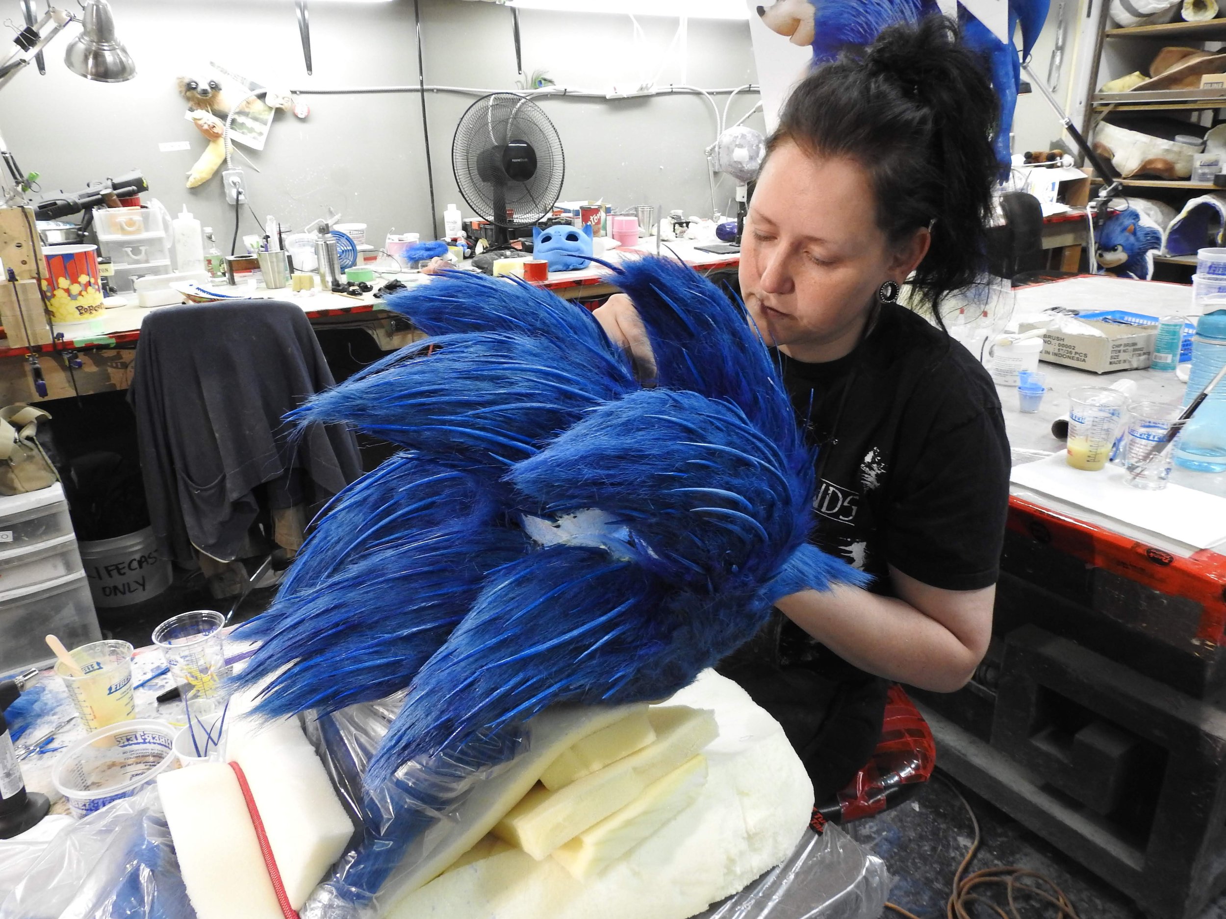 A woman working on a blue feathered creature puppet in a workshop filled with tools, supplies, and additional puppets.